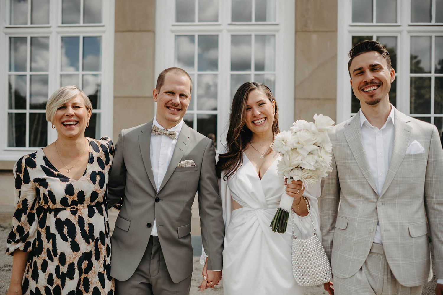 Wedding party in gray and white formal attire poses together outdoors in front of windows.