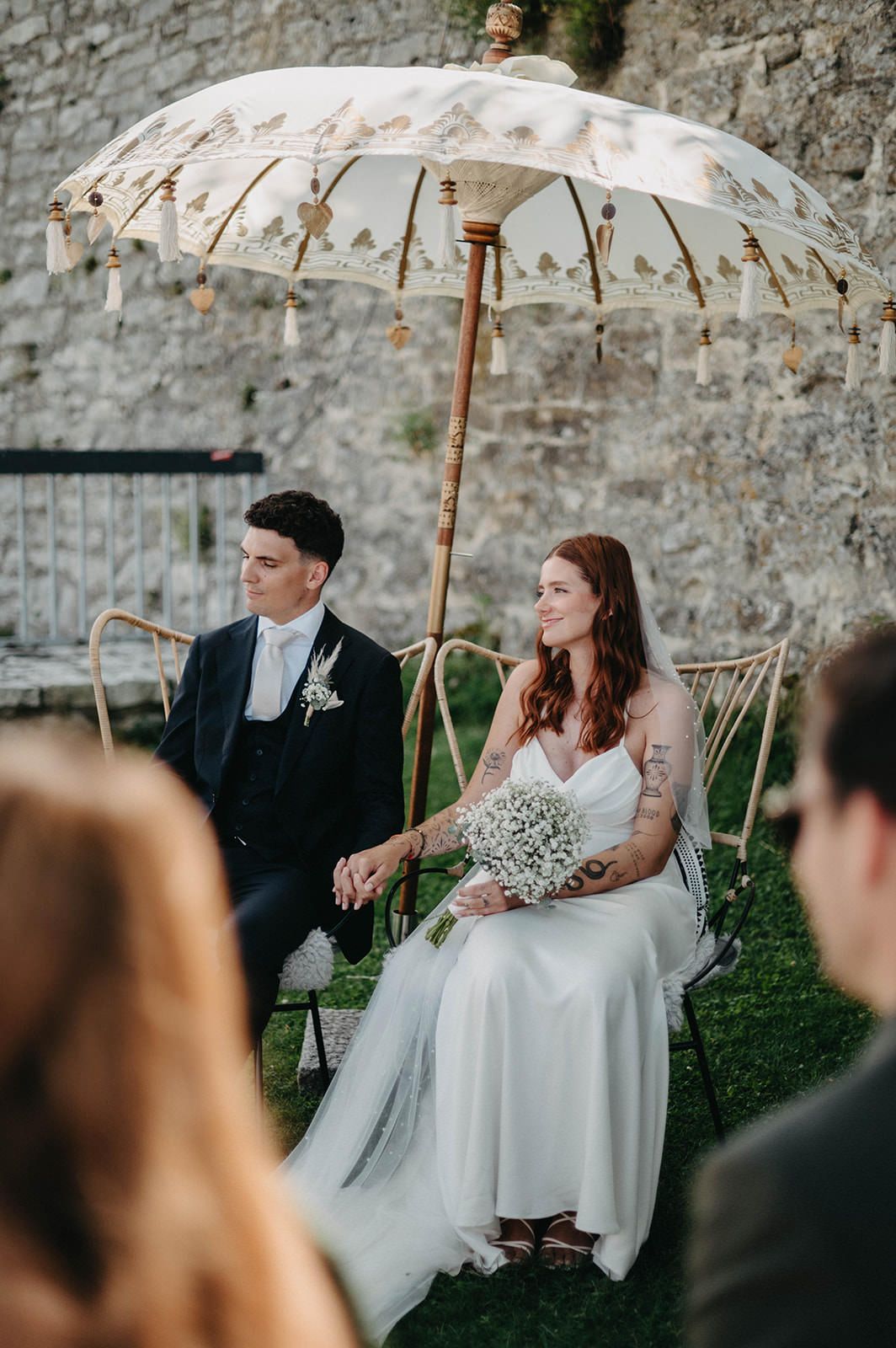 A couple sits outdoors under decorative parasols at their wedding reception with a stone wall backdrop.