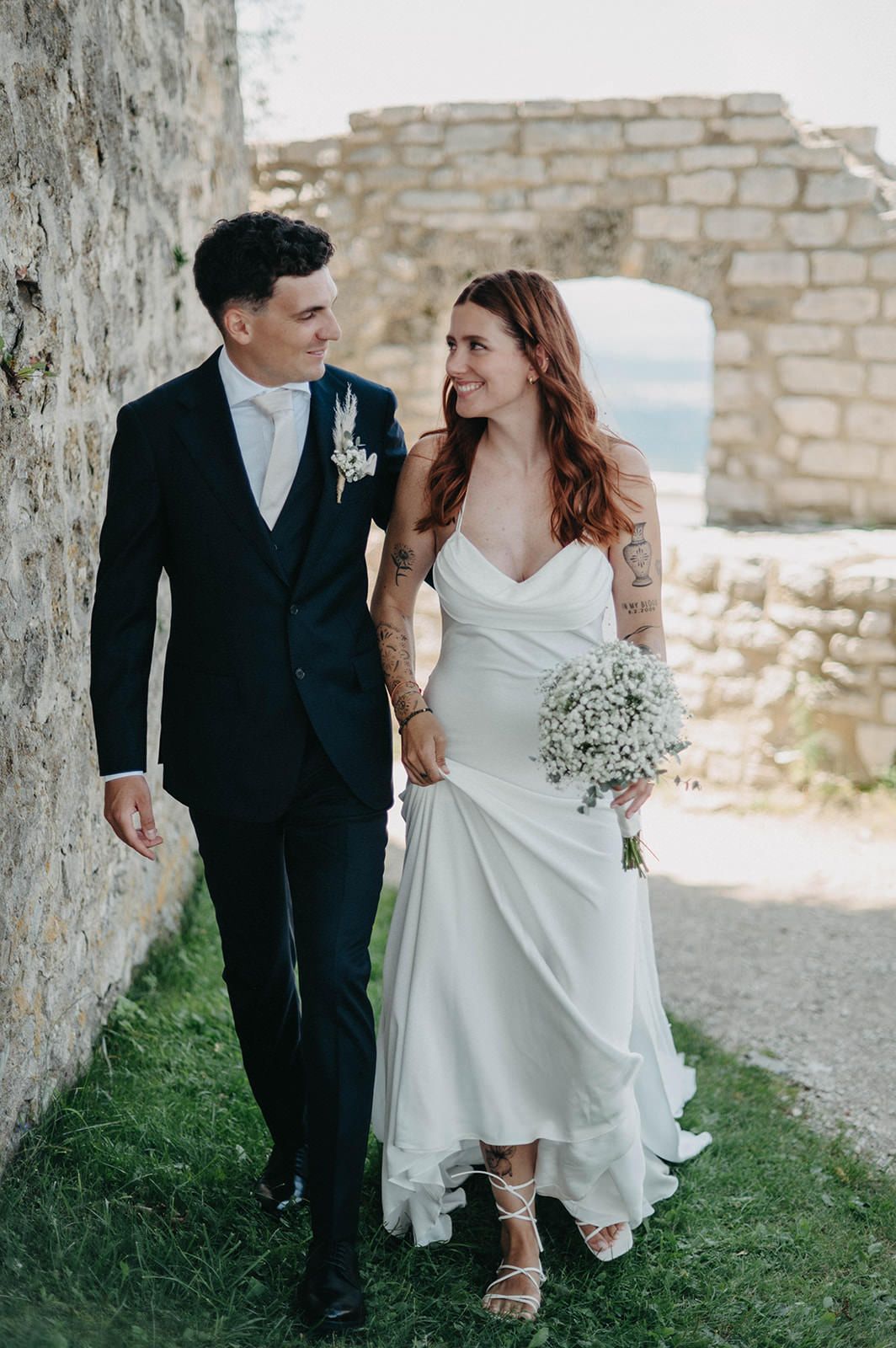 Romantic wedding couple walking together along stone walls and grass in casual elegant attire.