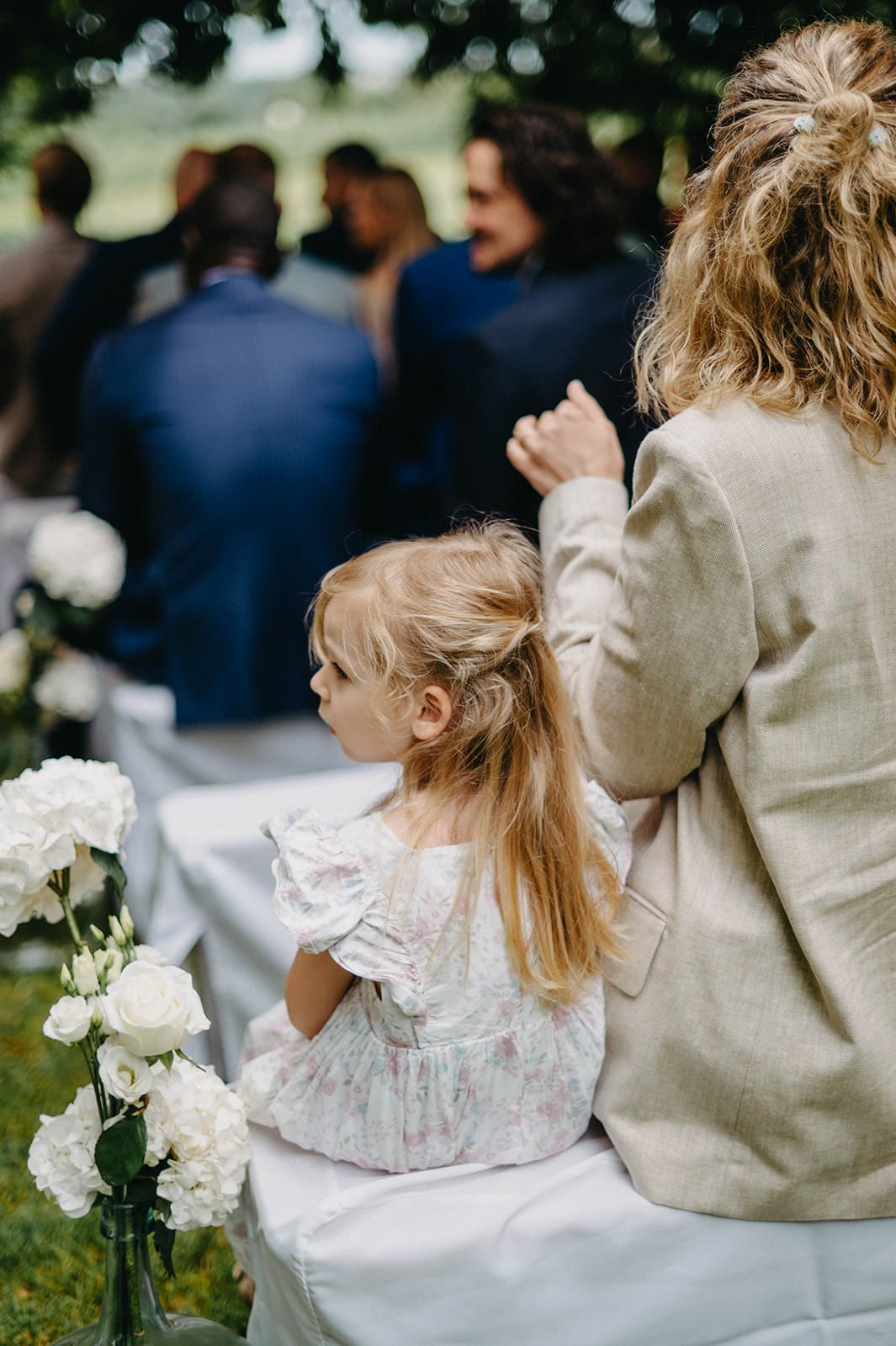 Child in white lace dress sits on blanket during outdoor wedding celebration with floral decorations.