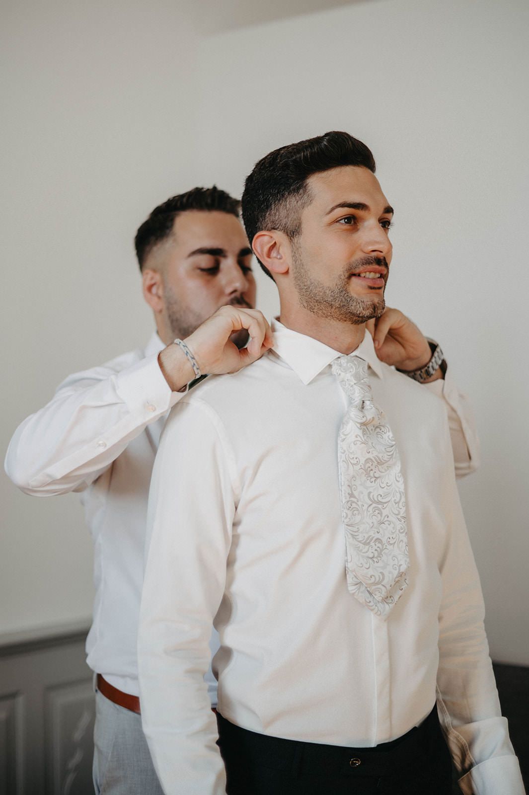 A groomsman helps adjust a white dress shirt and decorative lace tie during wedding preparations.