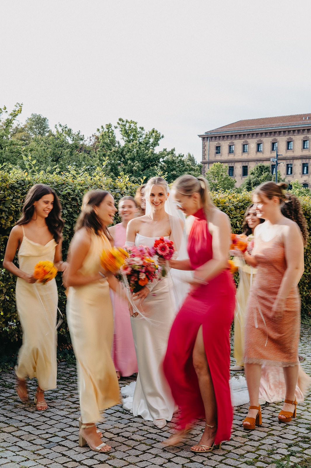 A bridal party in colorful dresses laughing together while standing on cobblestone pavement at a wedding celebration.