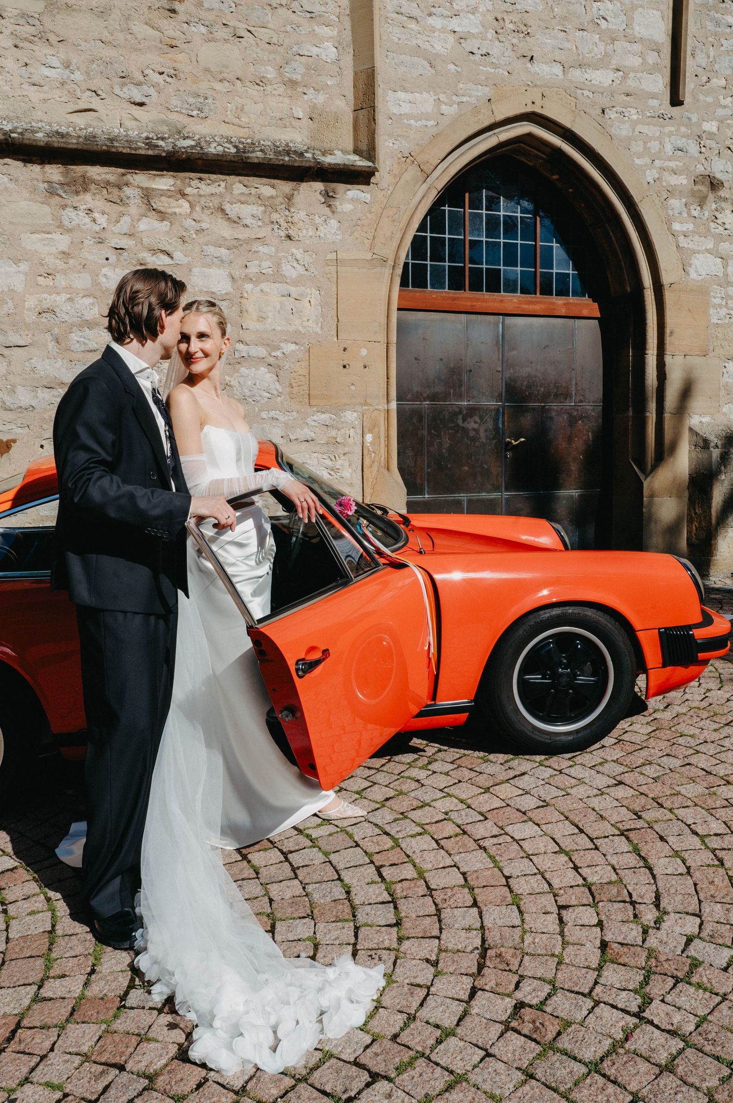 A couple in wedding attire poses next to a bright orange vintage sports car on a cobblestone courtyard.