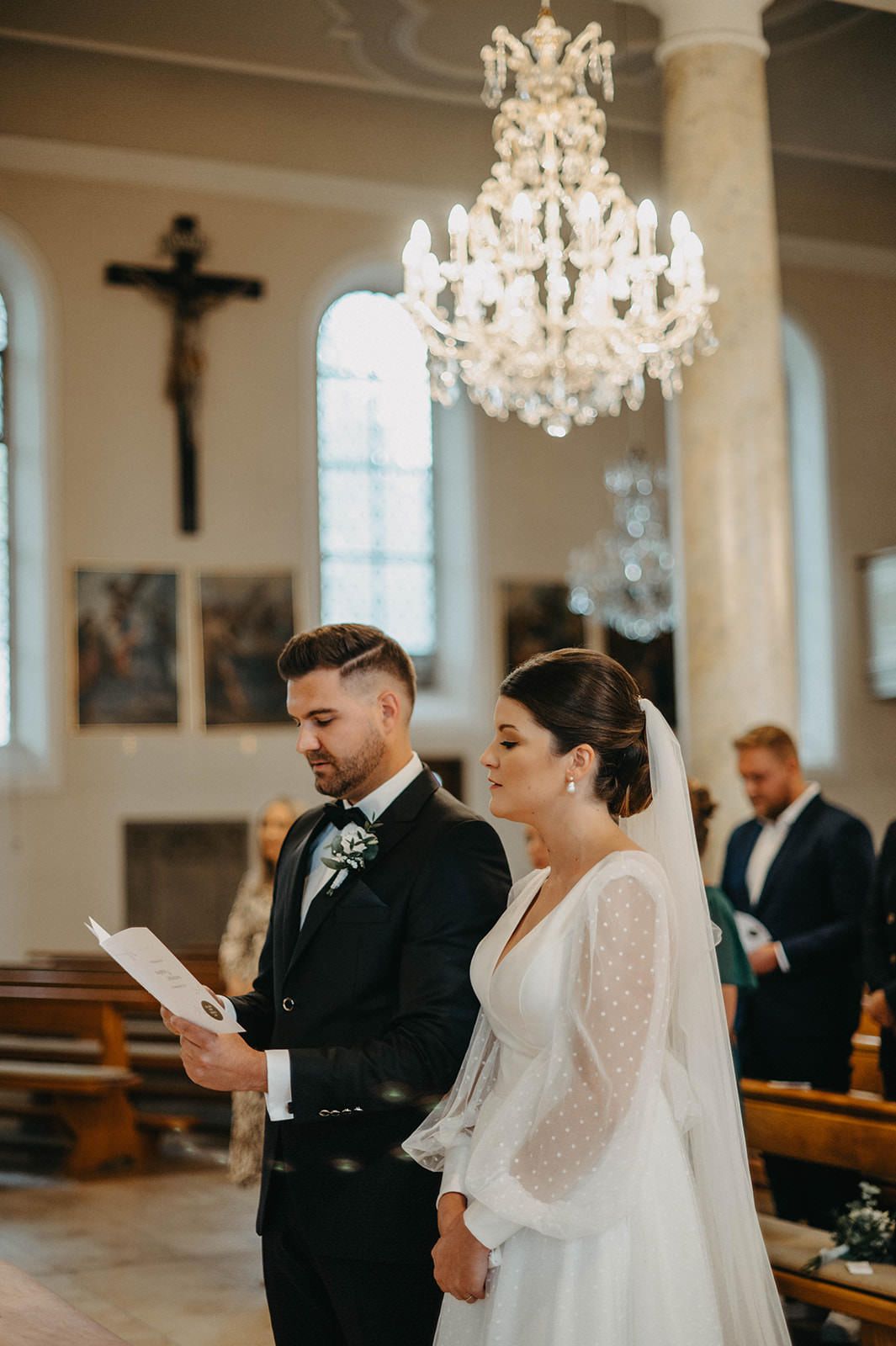 Couple exchanging vows during intimate church wedding ceremony beneath crystal chandeliers and religious decor.