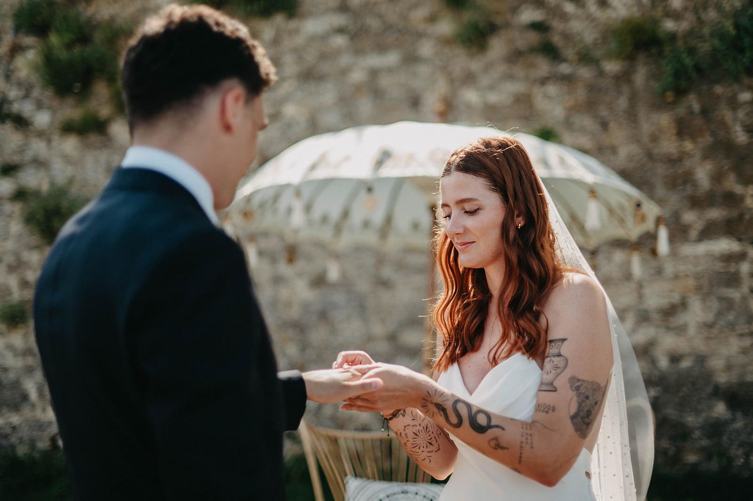 Couple shares intimate moments during outdoor wedding ceremony against stone wall backdrop.
