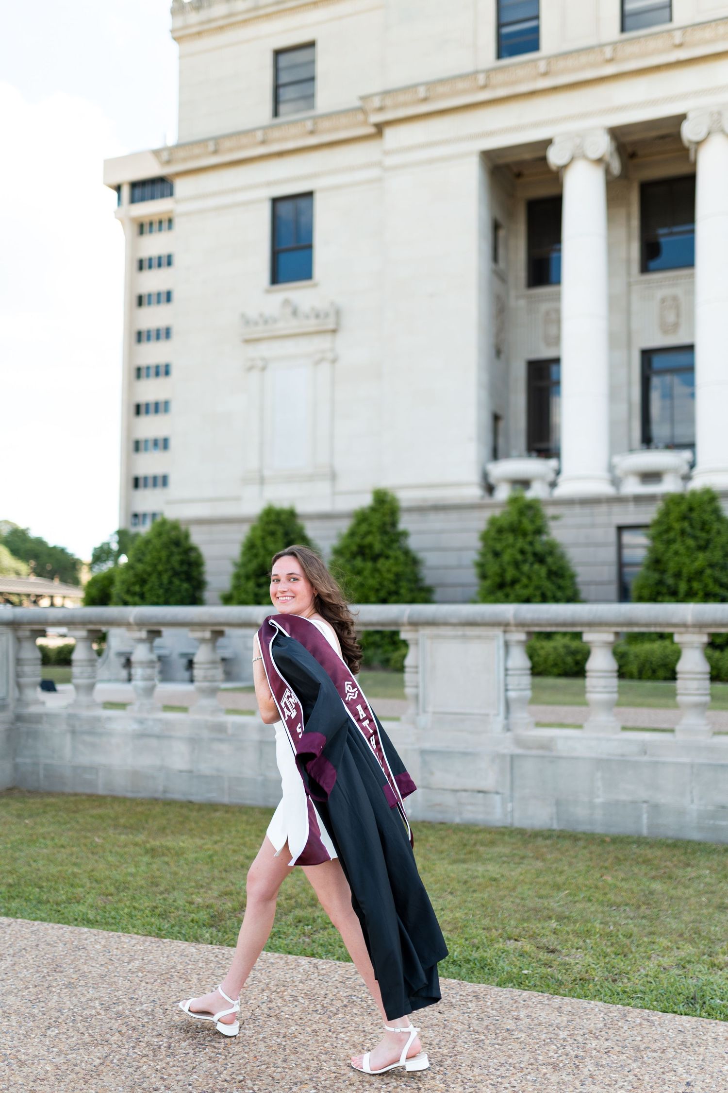 Smiling Texas A&M graduate walks in black graduation robe and maroon stole before neoclassical campus building. Grad Photos at the Admin