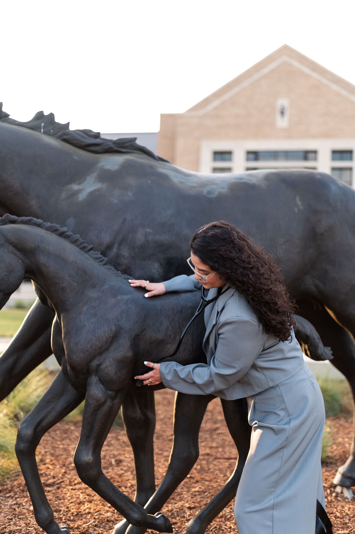 Curly-haired woman in gray suit uses stethoscope on bronze horse sculpture at Texas campus.