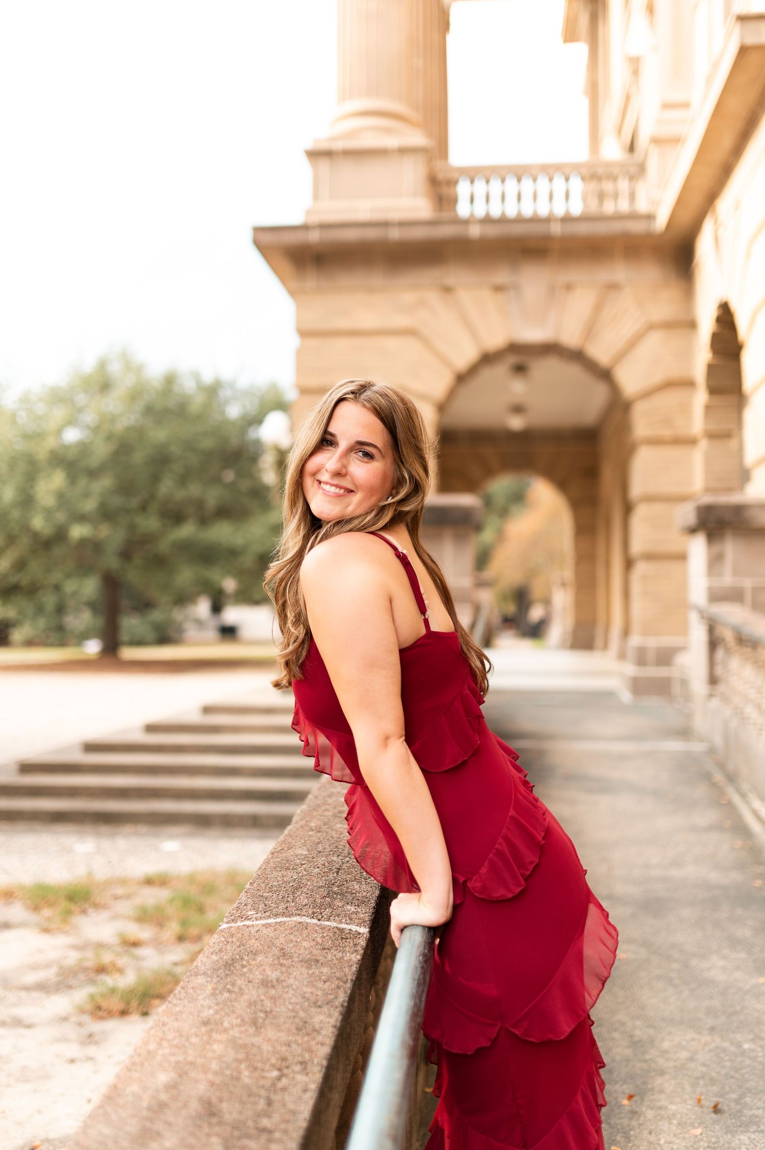 Woman in red ruffled dress smiles over shoulder while leaning on railing outside.