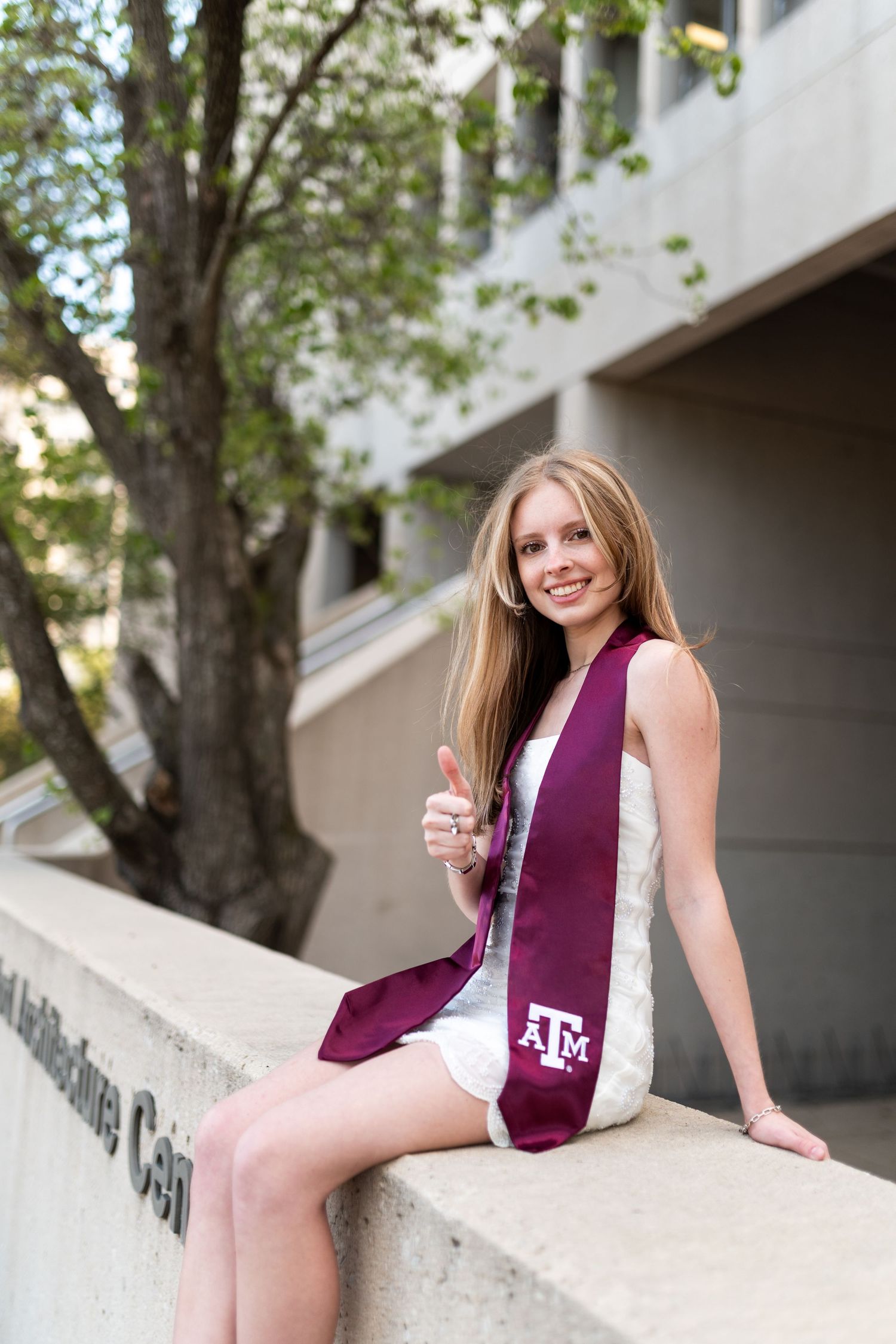 Smiling Texas A&M graduate in white dress with maroon stole gives thumbs up while sitting on concrete wall.
