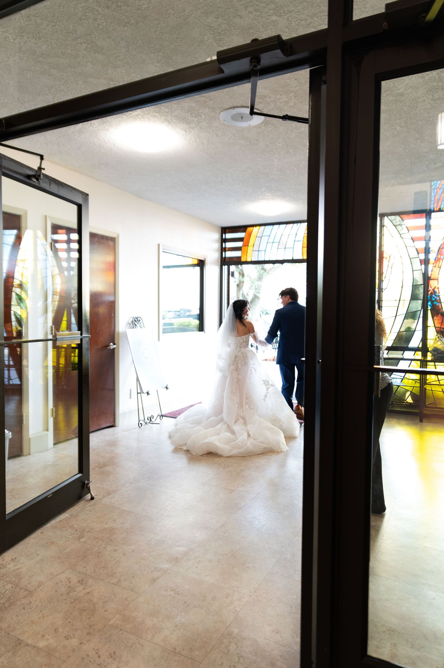 Bride in flowing white gown and groom stand in church foyer near stained glass windows before ceremony.