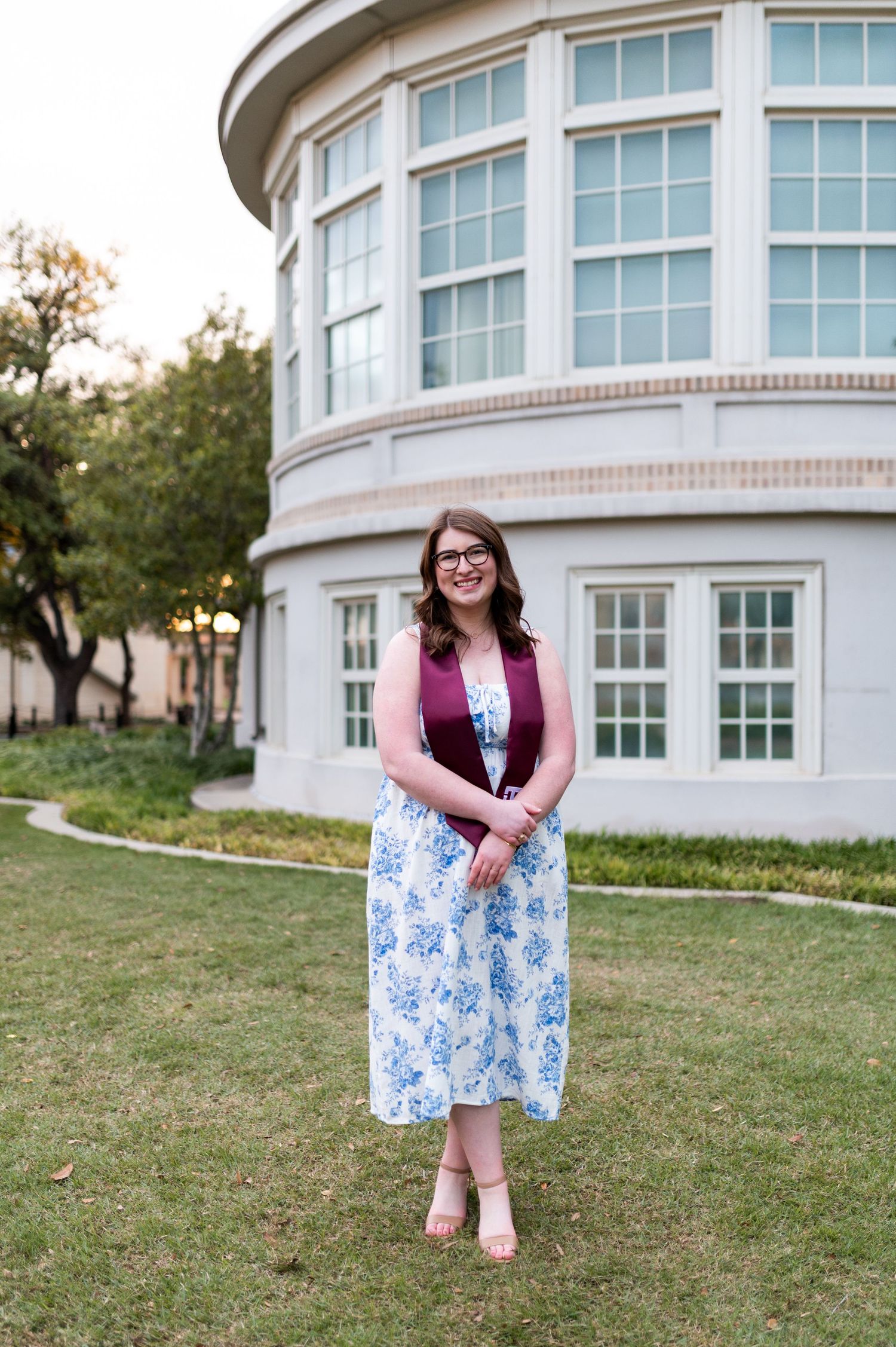 Texas A&M graduate posing in floral dress and maroon vest in front of a large circular building.