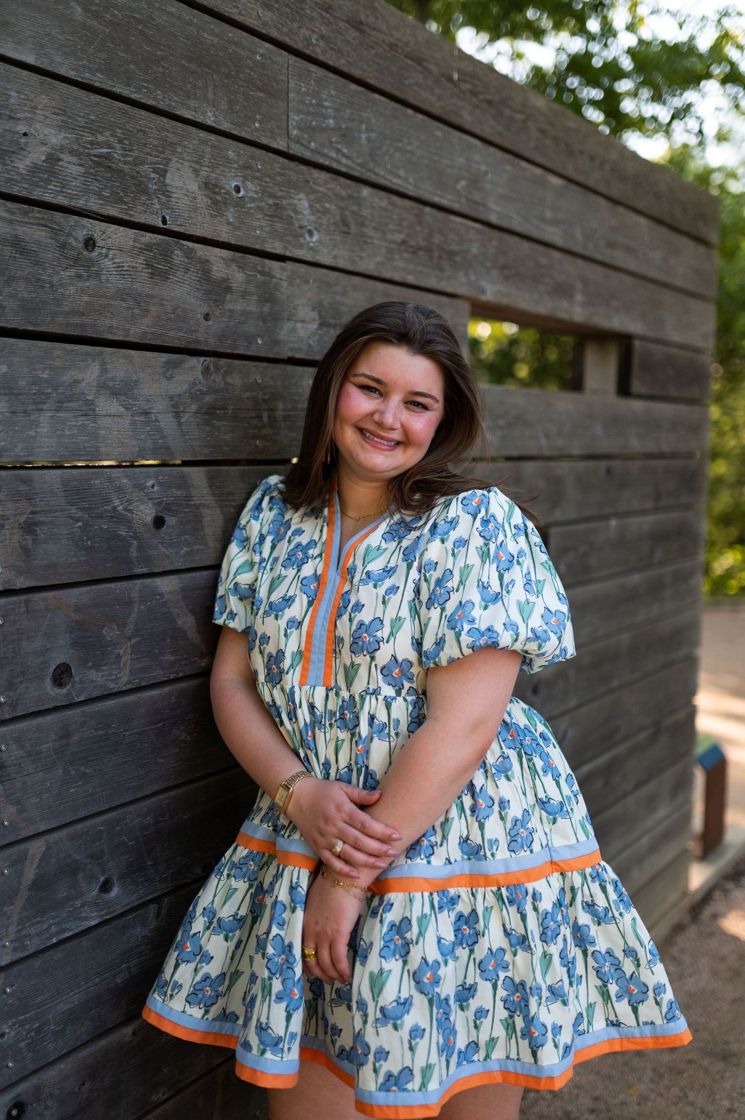 Smiling woman in blue floral dress with orange trim leaning against rustic wooden wall.