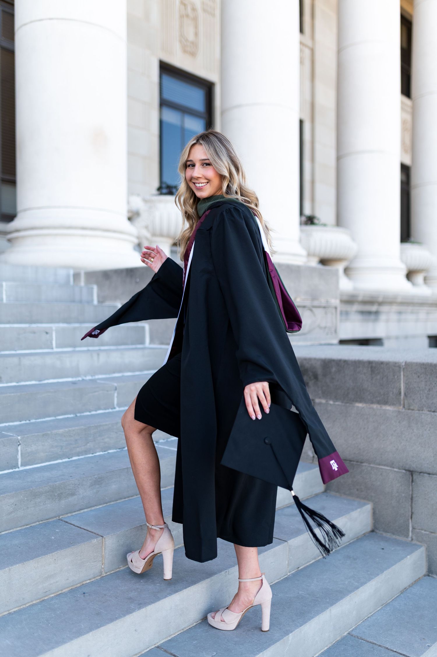 Smiling blonde woman in black graduation gown and heels posing on steps of Texas A&M University building. Texas A&M Grad Photos at the Admin