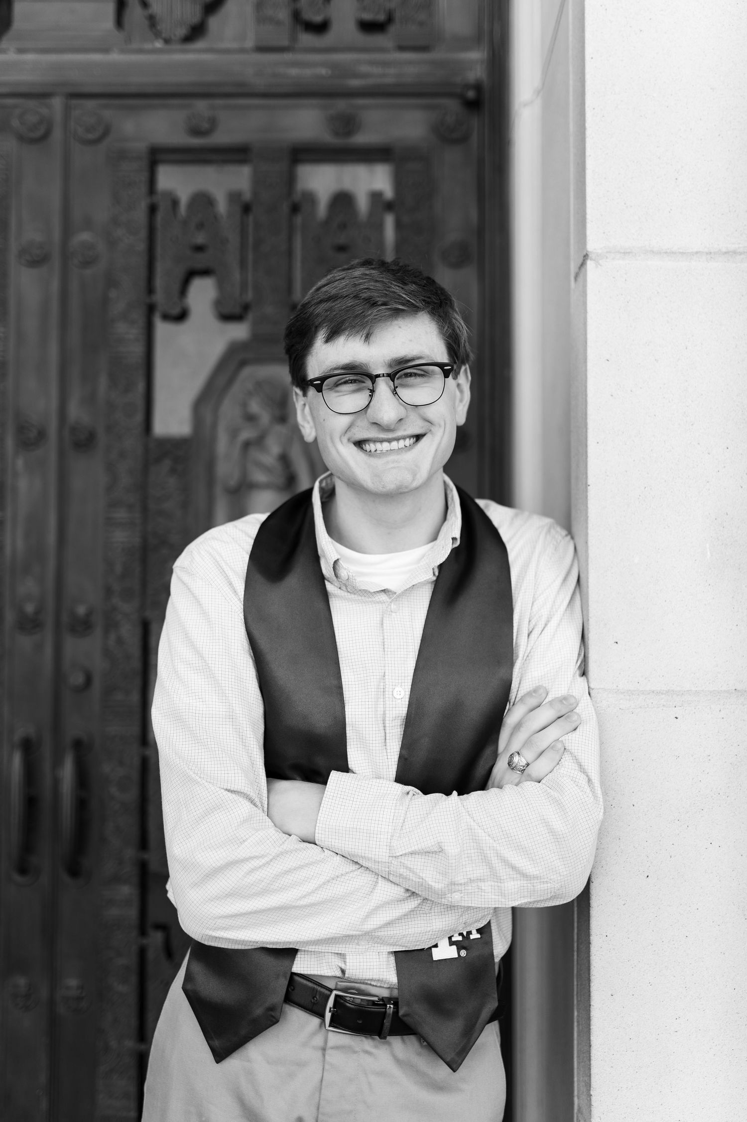 Black and white photo of smiling young man in glasses and vest leaning against ornate door. Texas A&M Grad Photos at the Admin building
