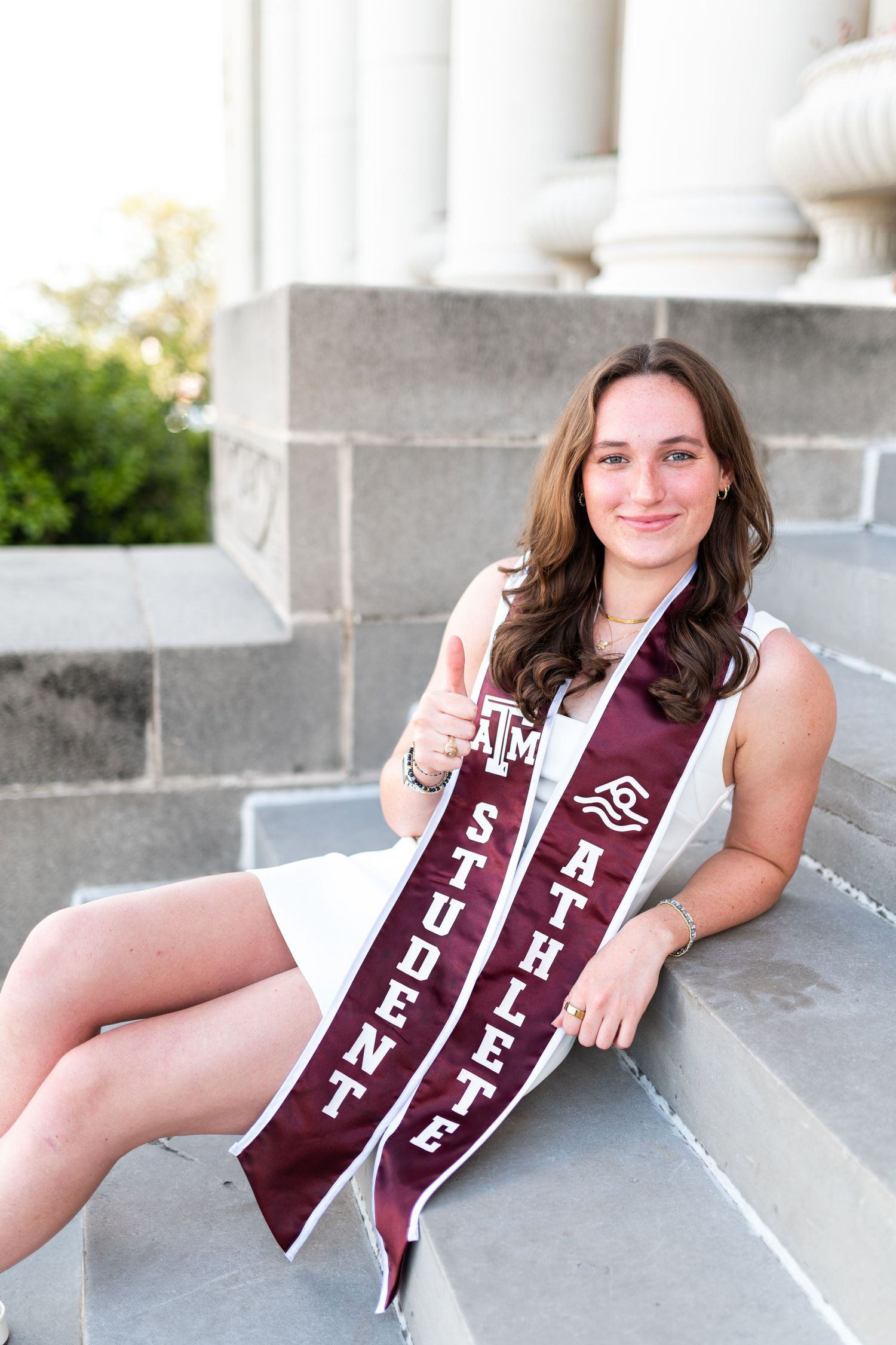 Texas A&M student-athlete graduate sits on steps displaying two maroon sashes with thumbs up gesture. Grad Photos at the Admin building
