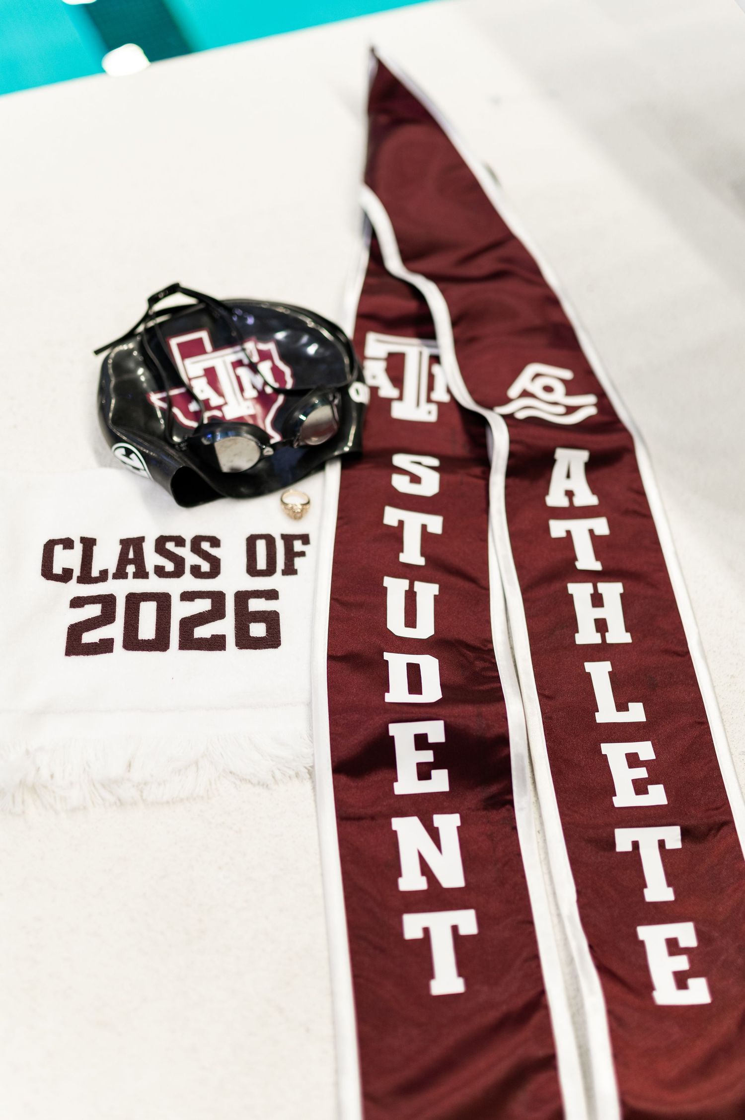 Texas A&M Class of 2026 student athlete graduation stole with swimming cap and ring displayed on white towel.