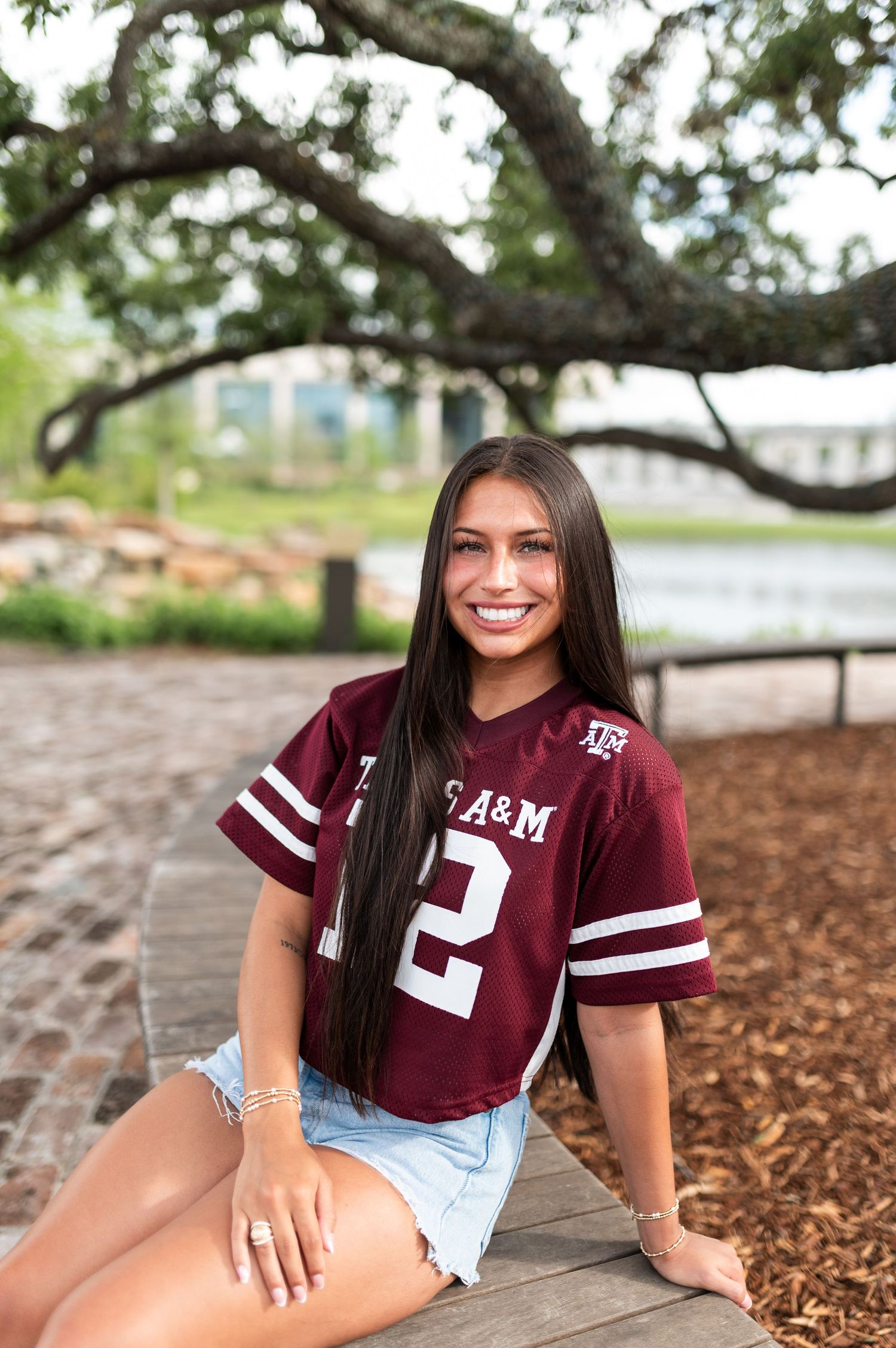 Young woman smiling in Texas A&M #12 maroon jersey and denim shorts sitting outdoors near a tree.