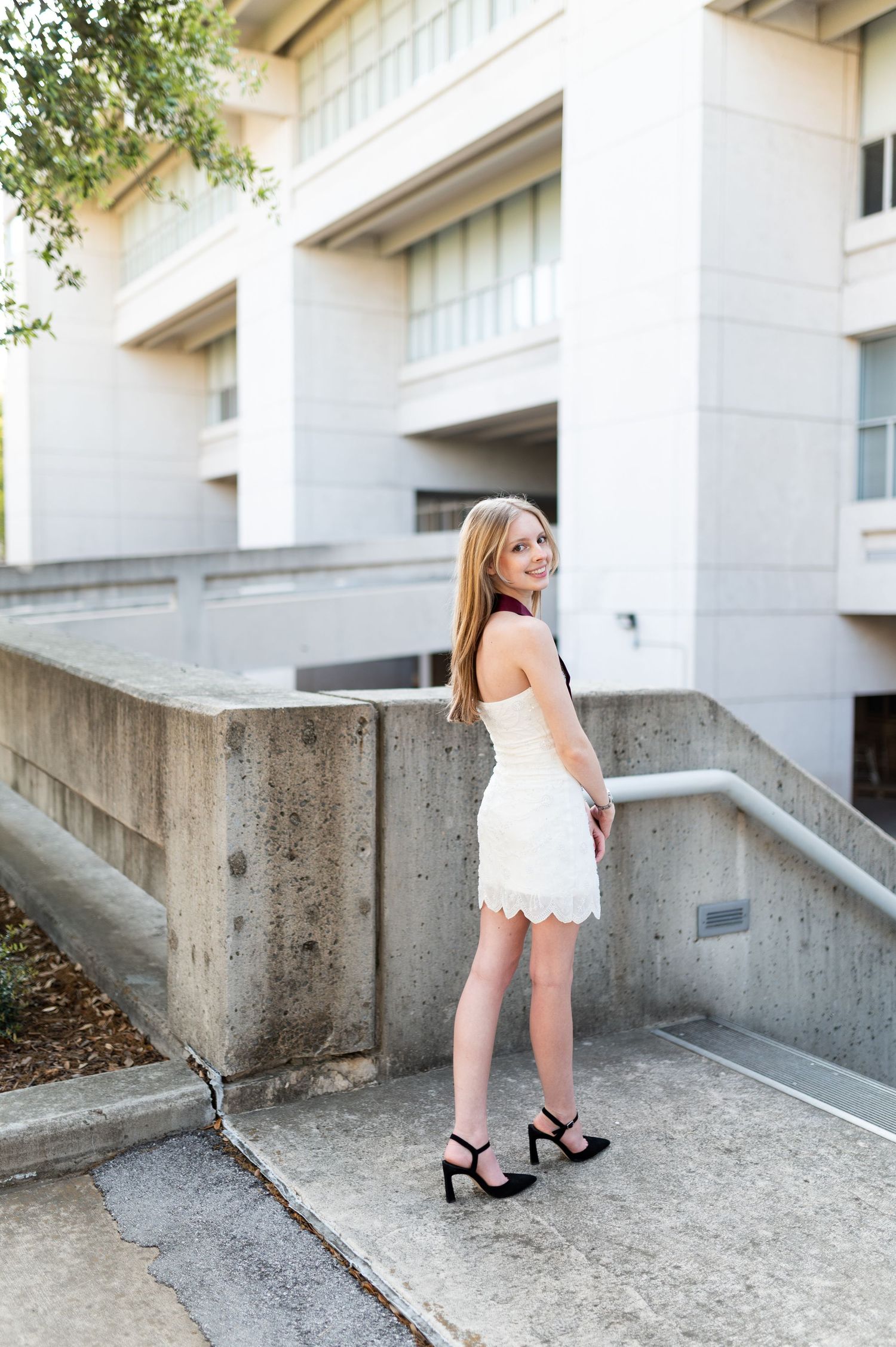 Blonde woman in white lace mini dress and black heels poses on concrete steps of modern campus building.