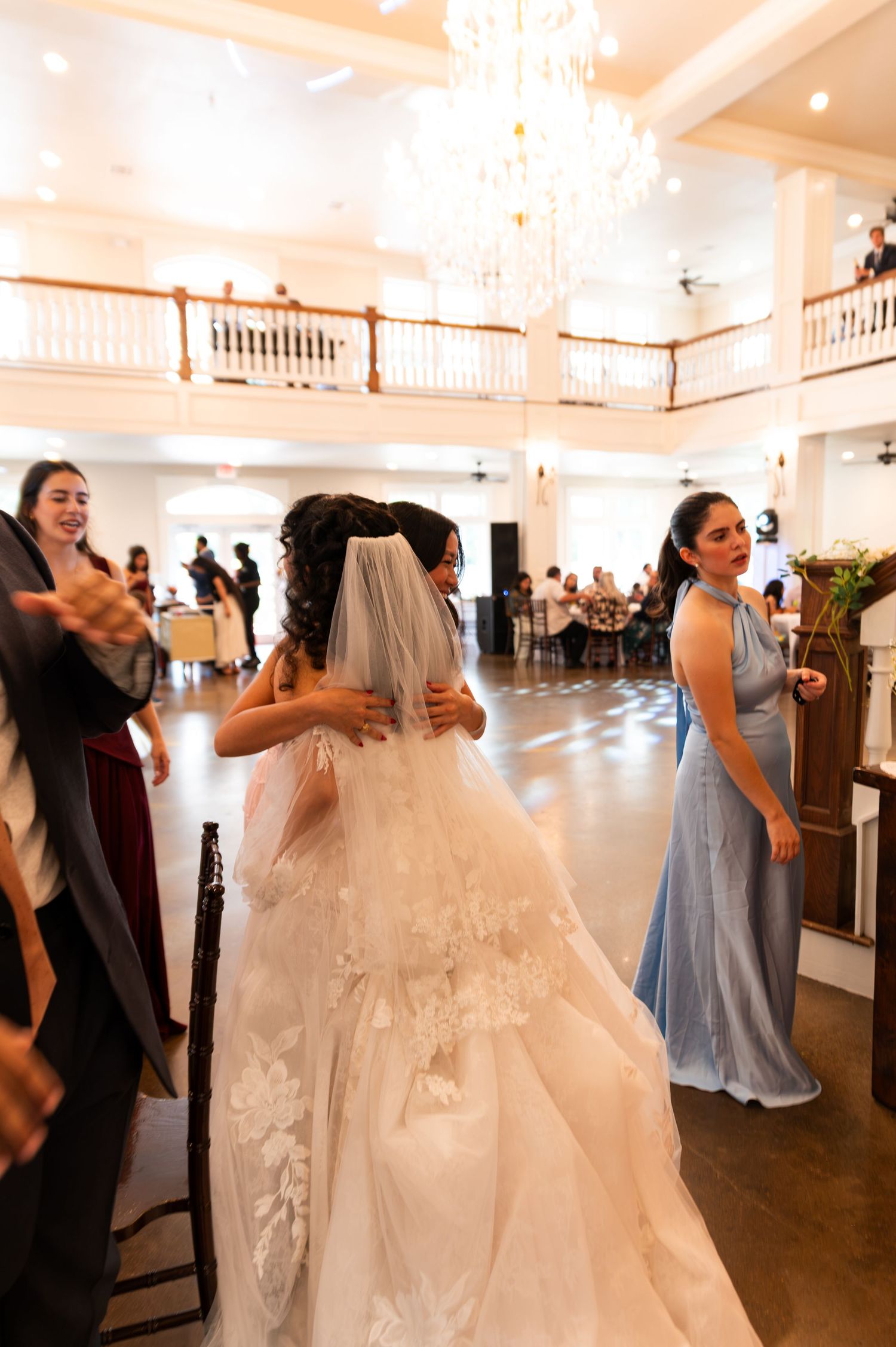 Bride in lace veil and gown hugs guest on dance floor at elegant wedding reception venue.