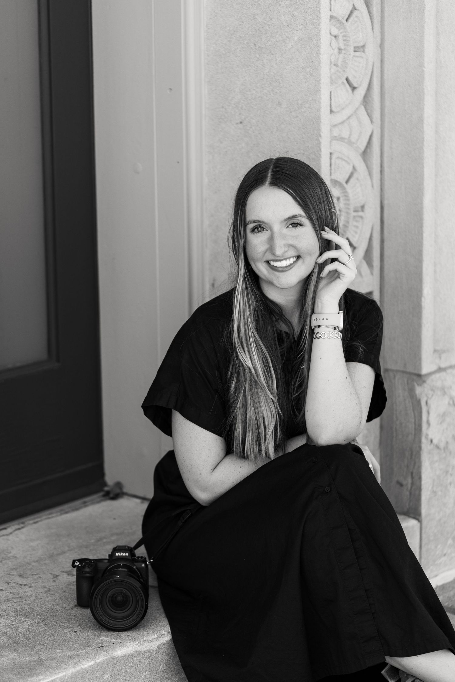 Black and white portrait of a smiling woman with long hair in a dark dress sitting on stone steps beside a camera.