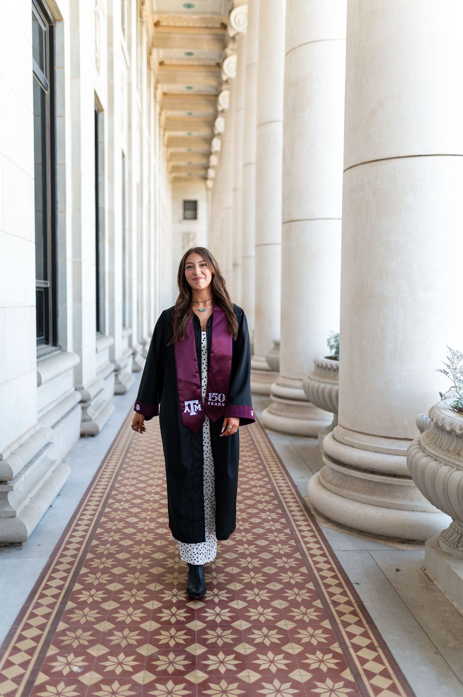 Female Texas A&M graduate walks through elegant columned hallway wearing black graduation robe and maroon stole. Grad Photos at the Admin