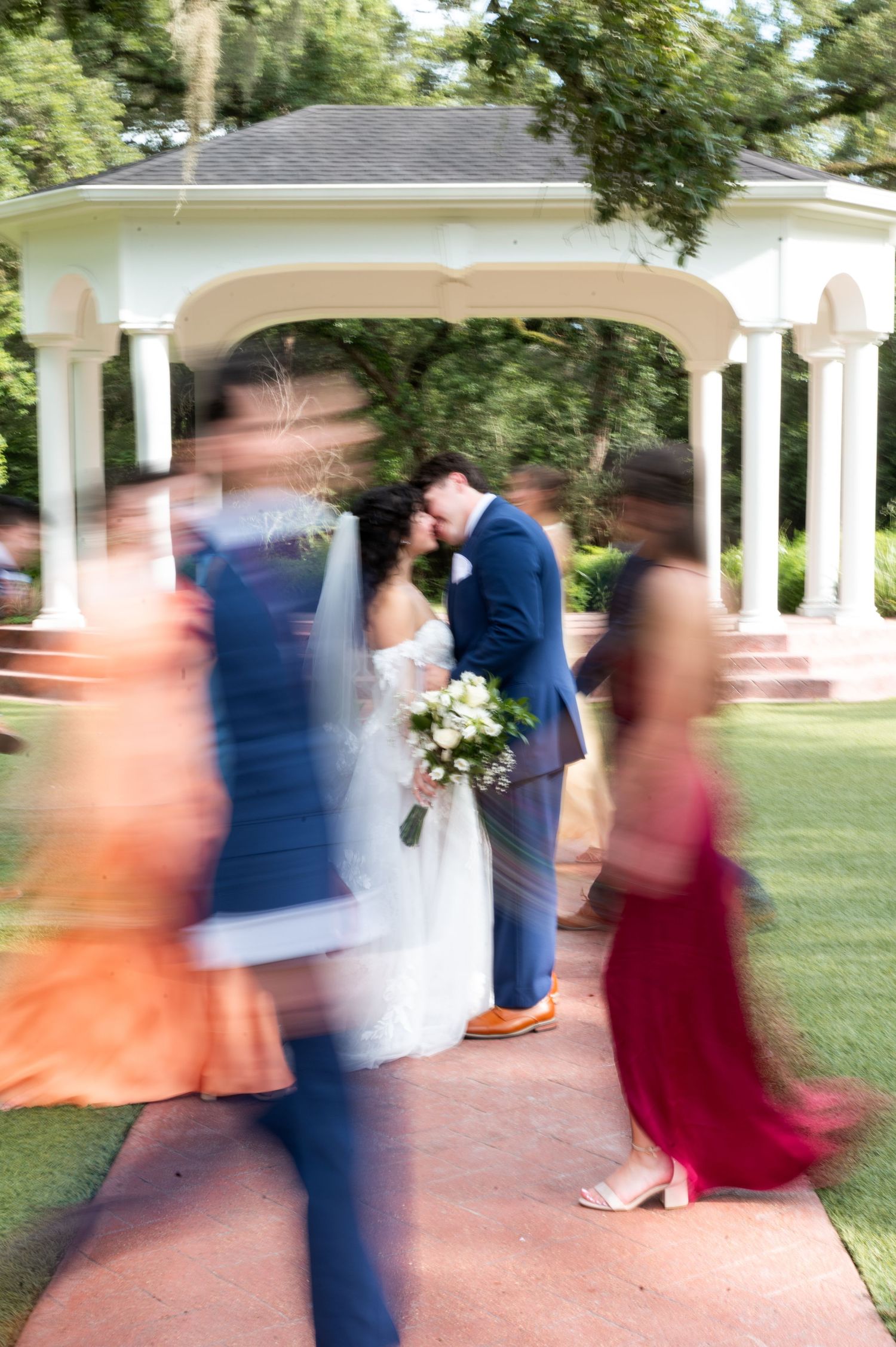 Bride and groom share a kiss under a white gazebo surrounded by blurred wedding guests in motion.