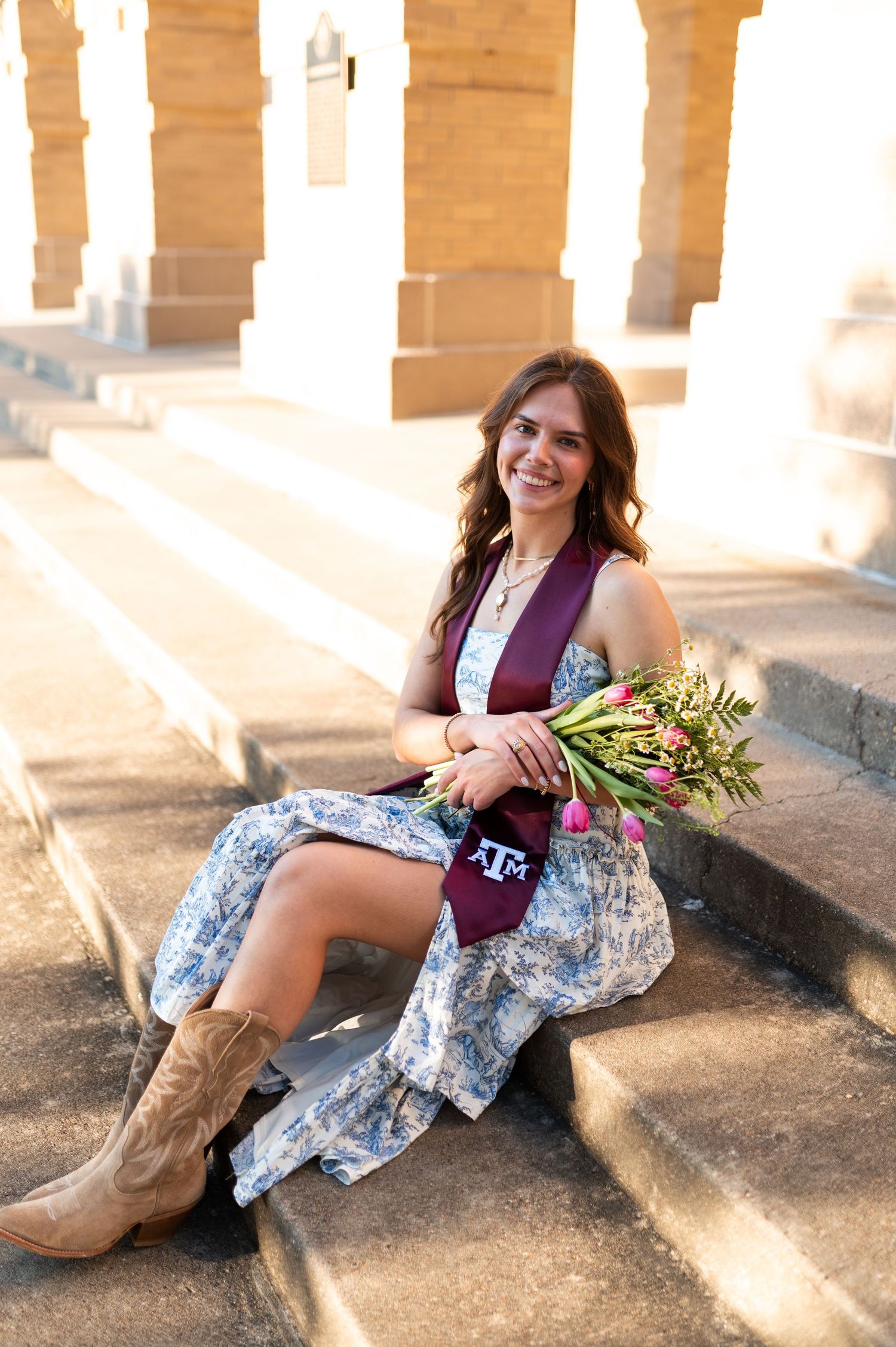Smiling female Texas A&M graduate sits on steps holding flowers wearing floral dress and maroon graduation stole.