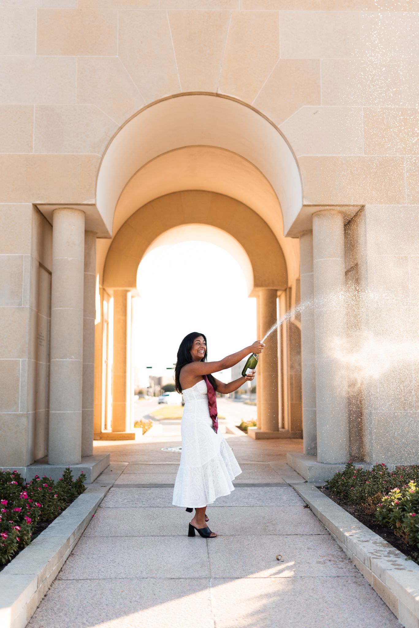 Woman in white dress popping champagne bottle under stone archway outdoors.