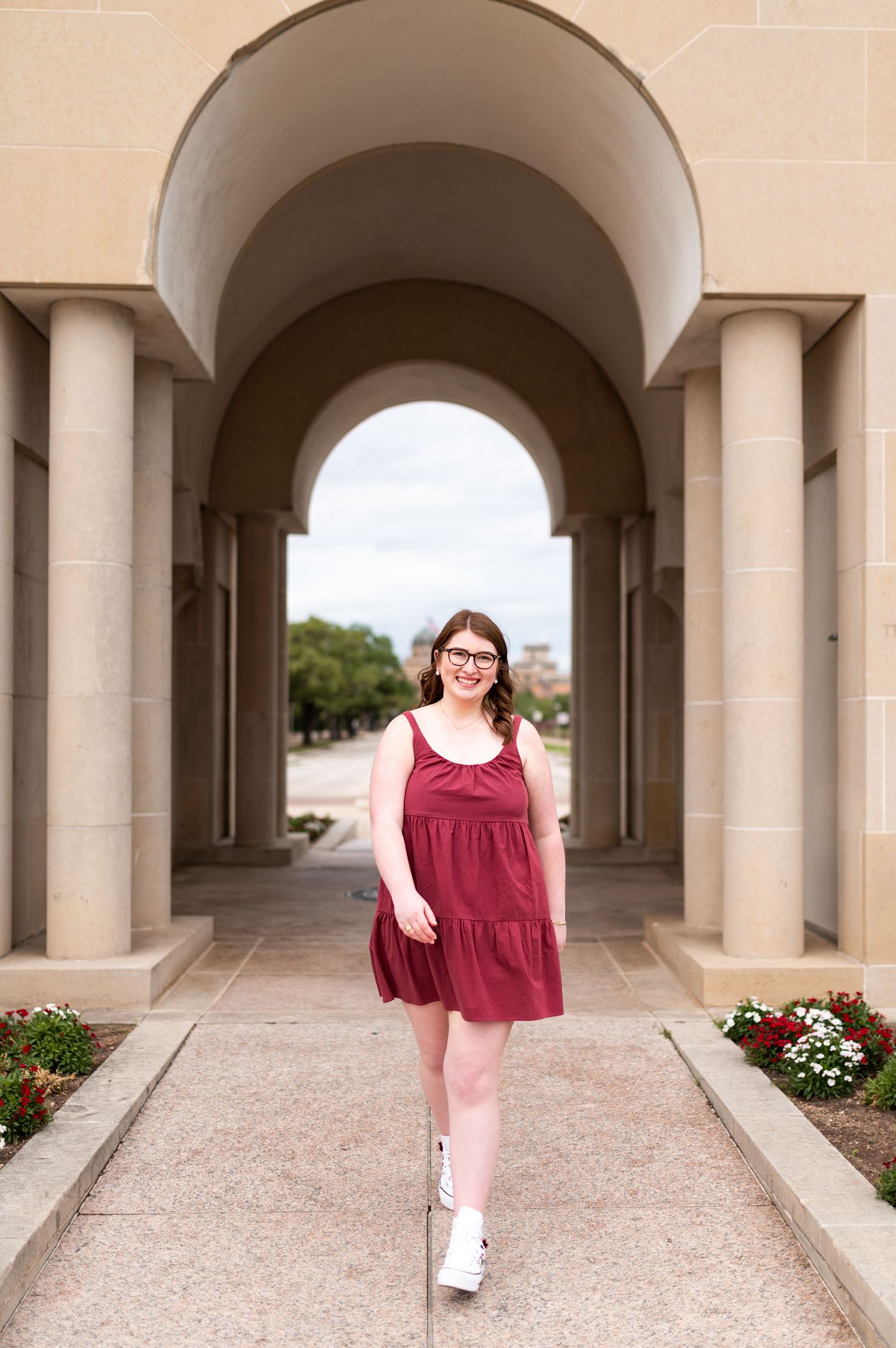 Texas A&M graduate in maroon dress walking confidently through stone archway on sunny campus.
