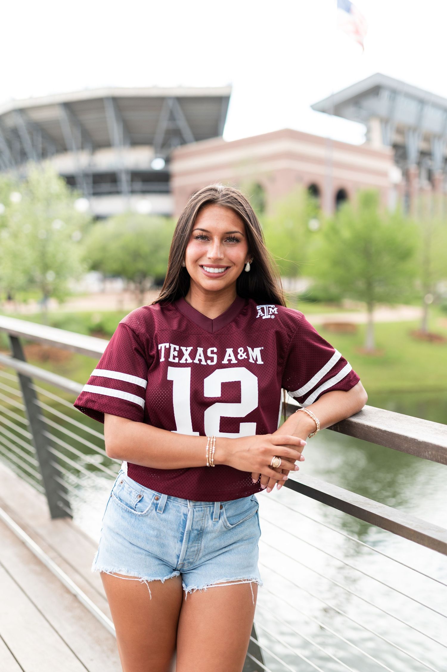 Smiling young woman in Texas A&M maroon number 12 jersey and denim shorts near Kyle Field stadium.