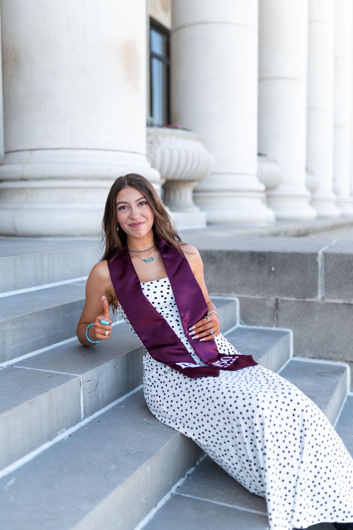 Texas A&M female graduate sits on stone steps in white polka dot dress with maroon graduation stole. Grad Photos at the Admin building