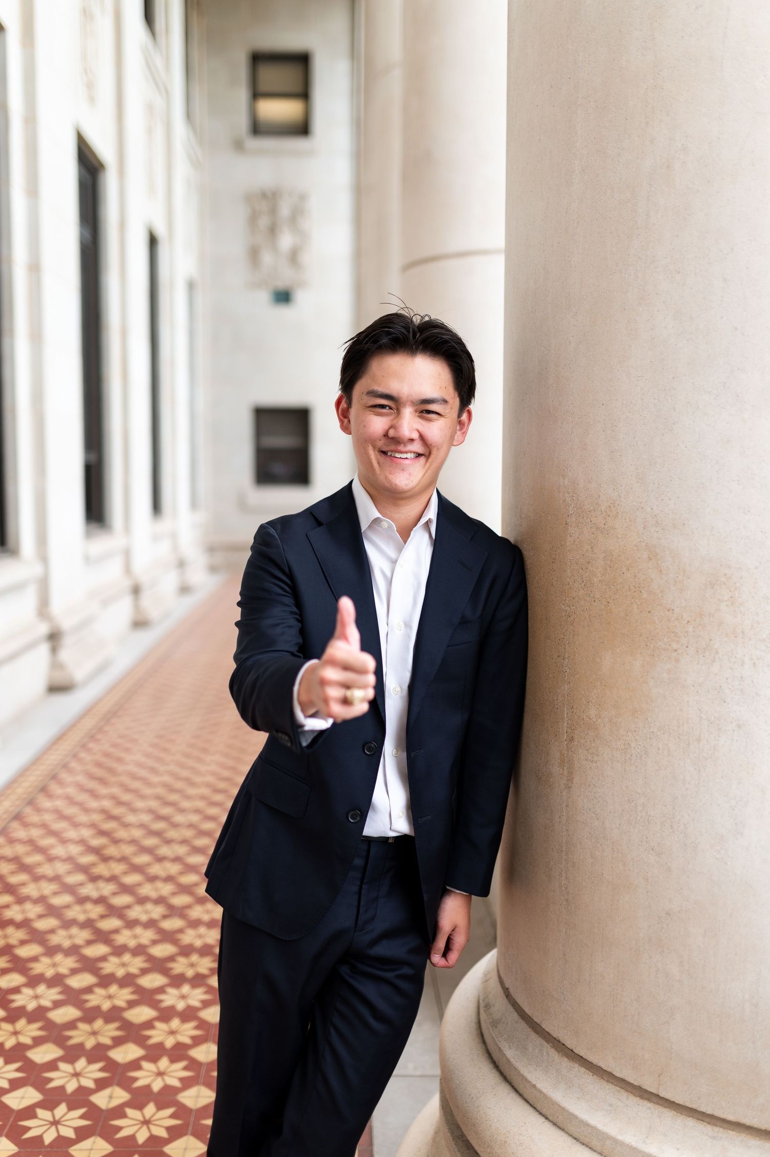 Smiling young man in navy suit gives thumbs up leaning against column in elegant hallway. Grad Photos at the Admin building