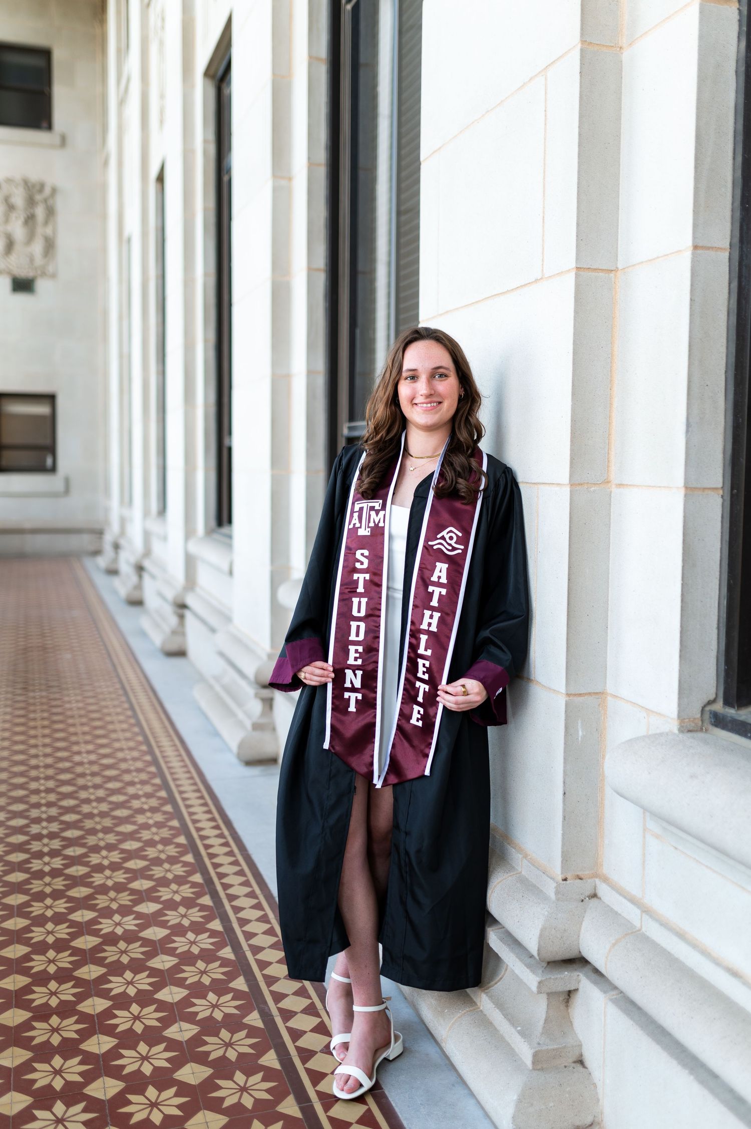 Texas A&M graduate in black cap and gown with student athlete stole poses in campus hallway. Grad Photos at the Admin building