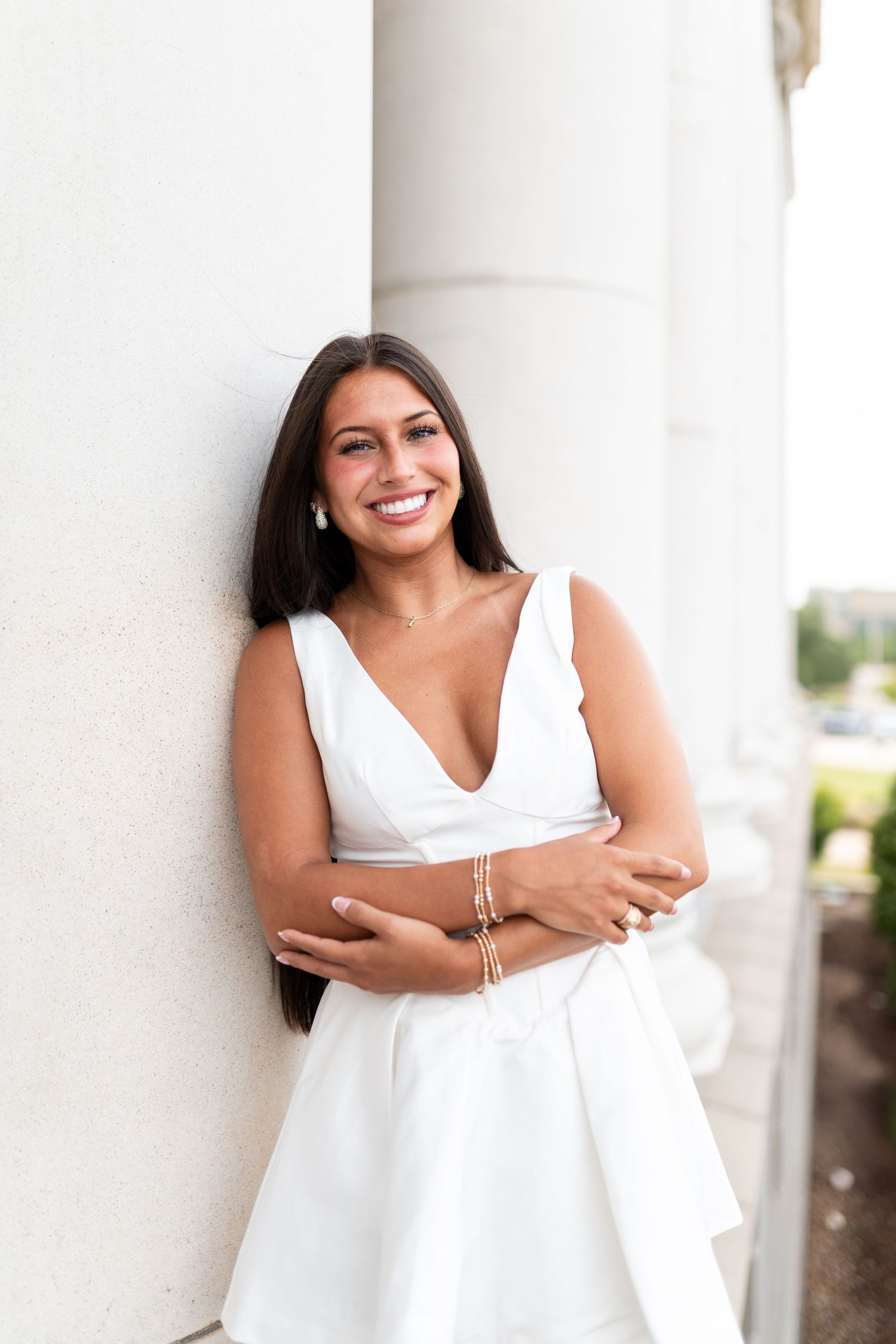 Smiling young woman in white dress leaning against white columns. Grad Photos at the Admin building