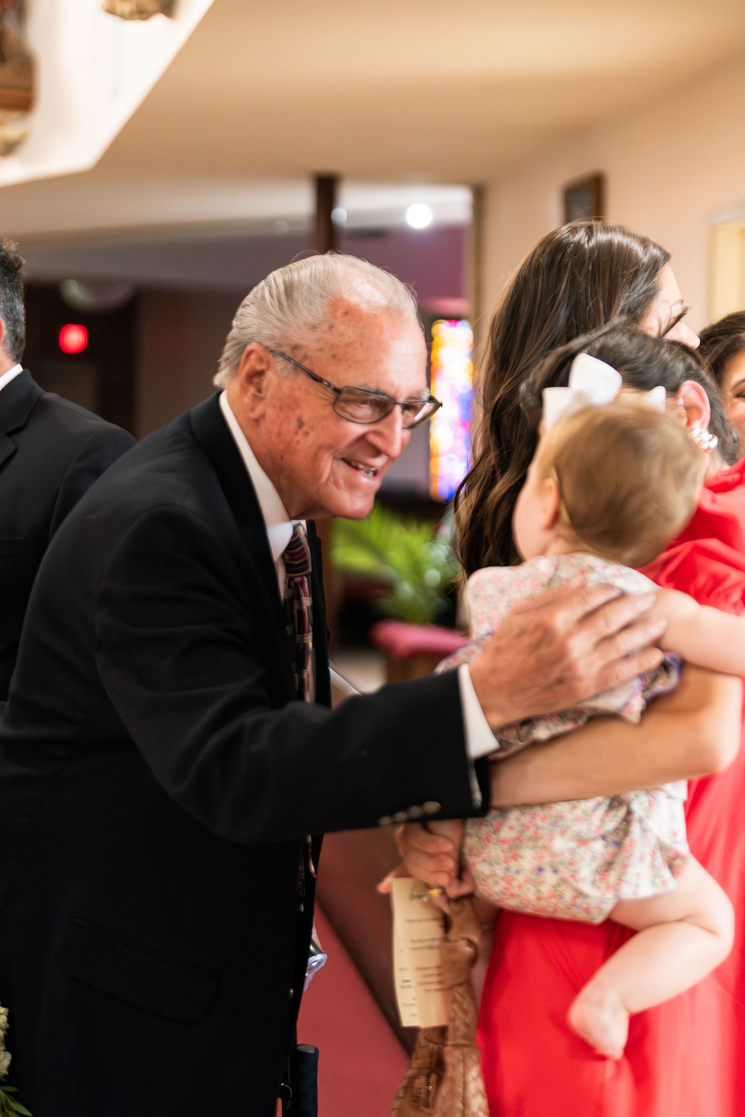 Elderly man in suit smiling warmly at baby held by woman in red dress at church.