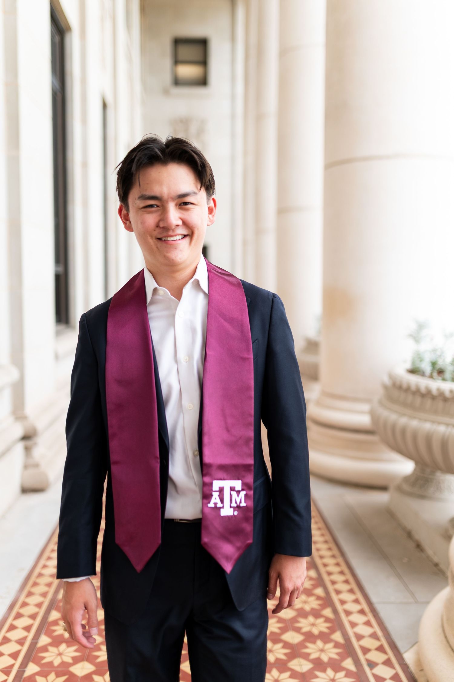 Smiling Texas A&M graduate in navy suit wearing maroon ATM graduation stole in campus hallway. Grad Photos at the Admin building