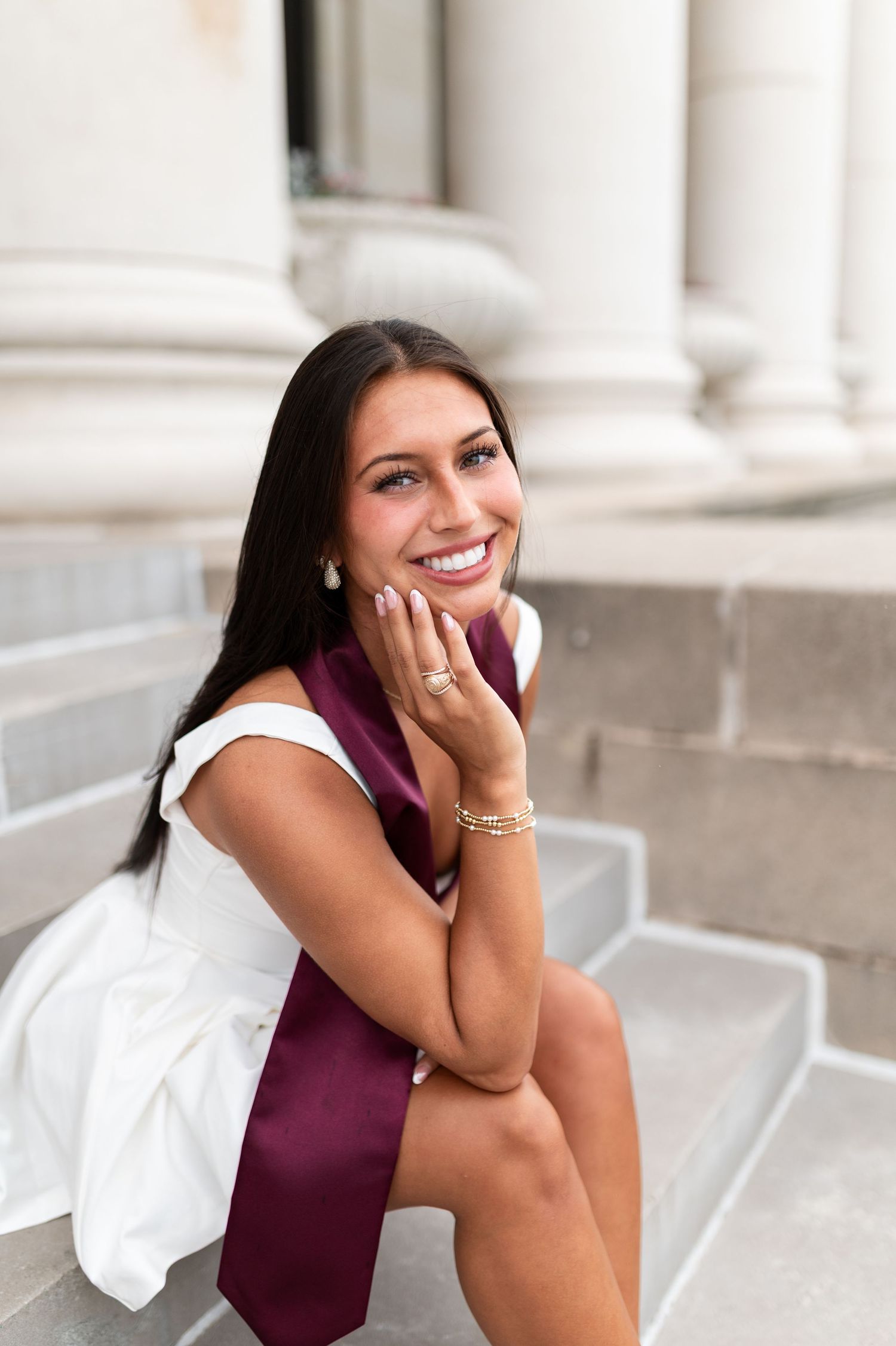Smiling Texas A&M graduate in white dress sitting on steps with maroon stole. Grad Photos at the Admin building