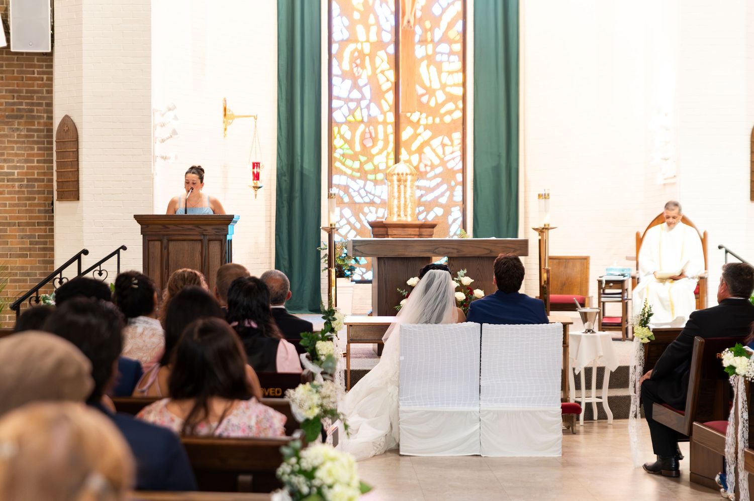 A bride and groom sit together during a church wedding ceremony with stained glass windows and guests seated in pews.