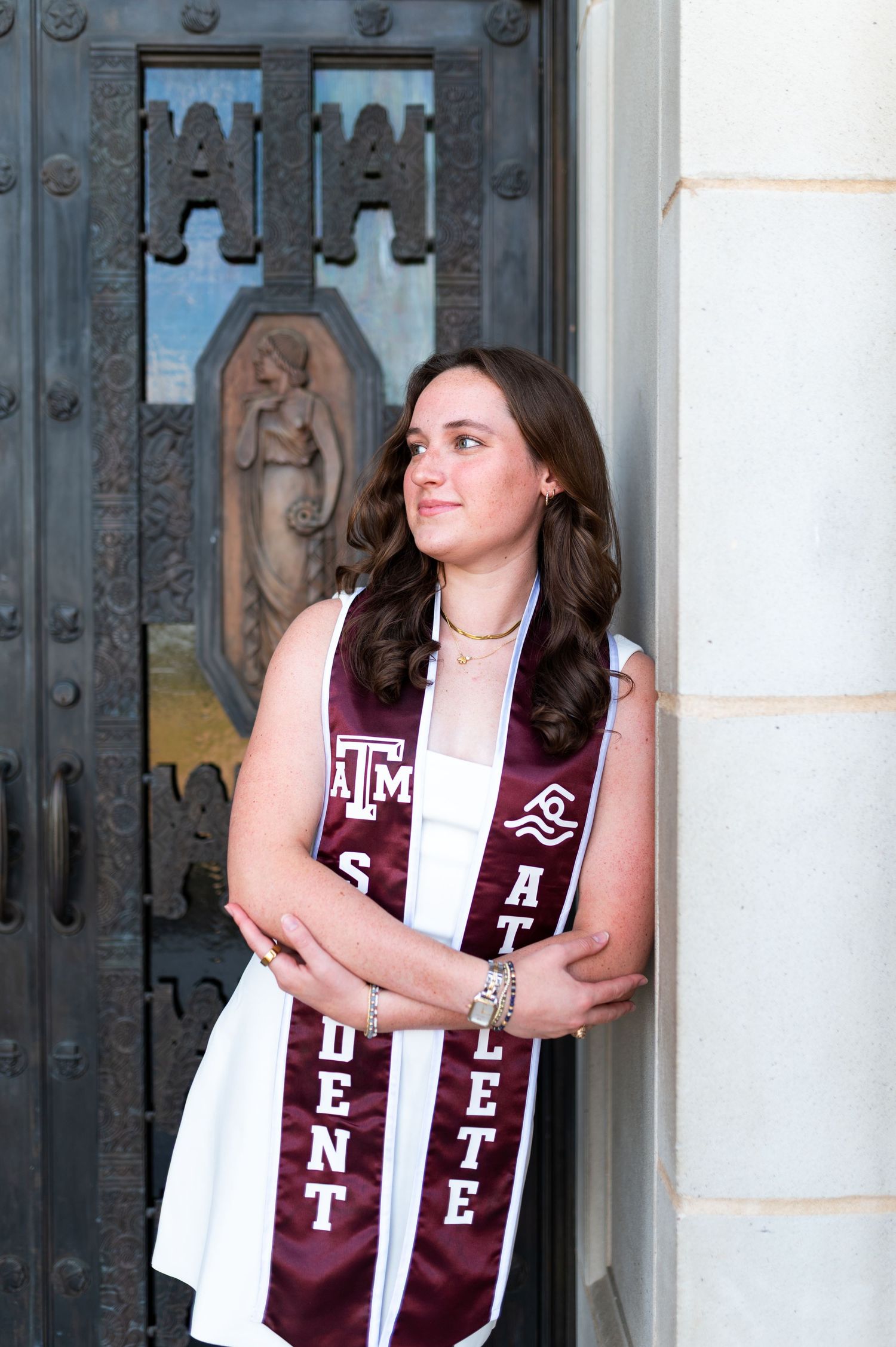 Texas A&M student athlete poses in white dress with maroon graduation stole at ornate campus doors. Grad Photos at the Admin building