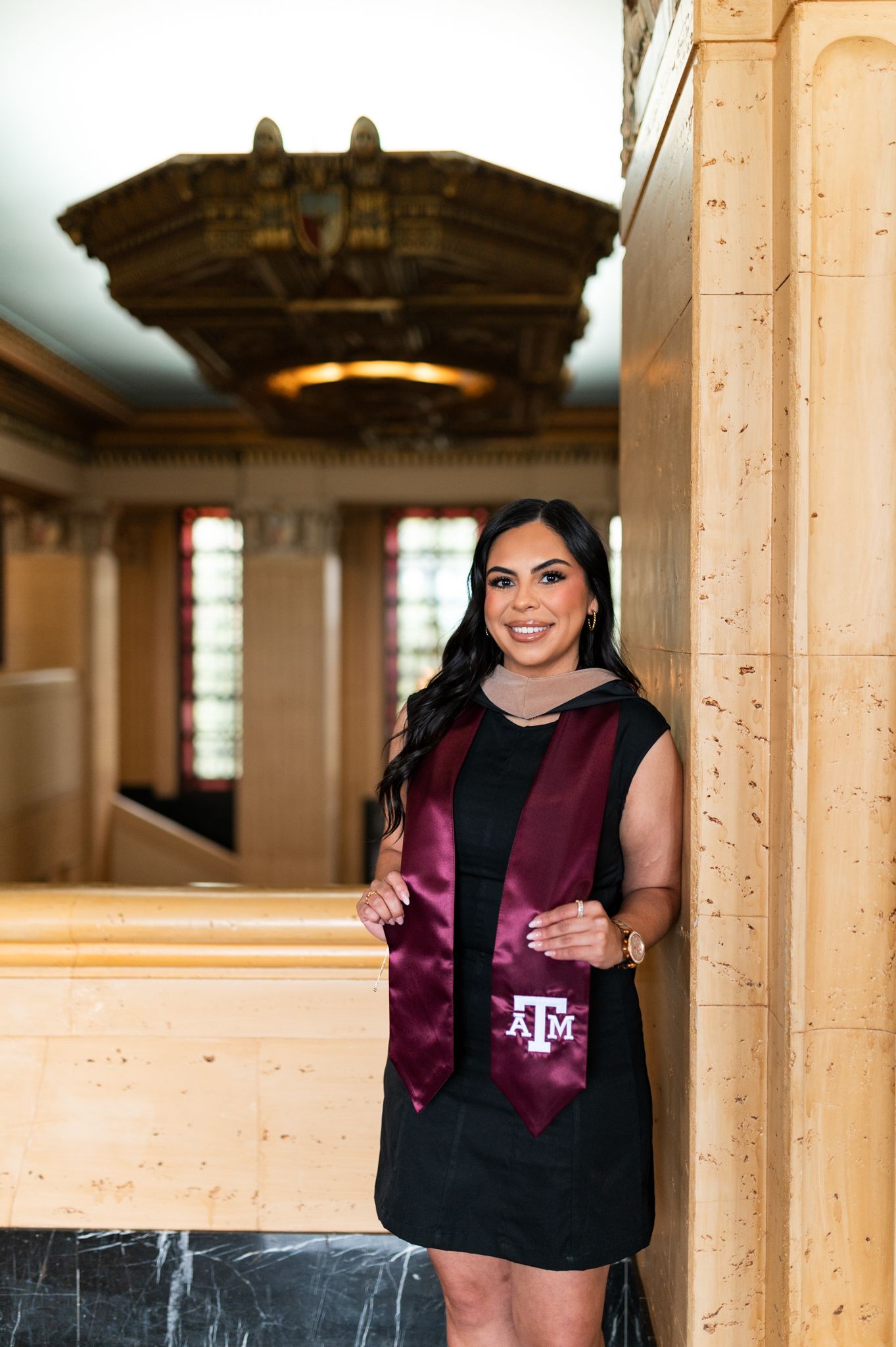 Smiling female Texas A&M graduate in black dress holding maroon ATM stole indoors. Grad Photos at the Texas A&M Admin building