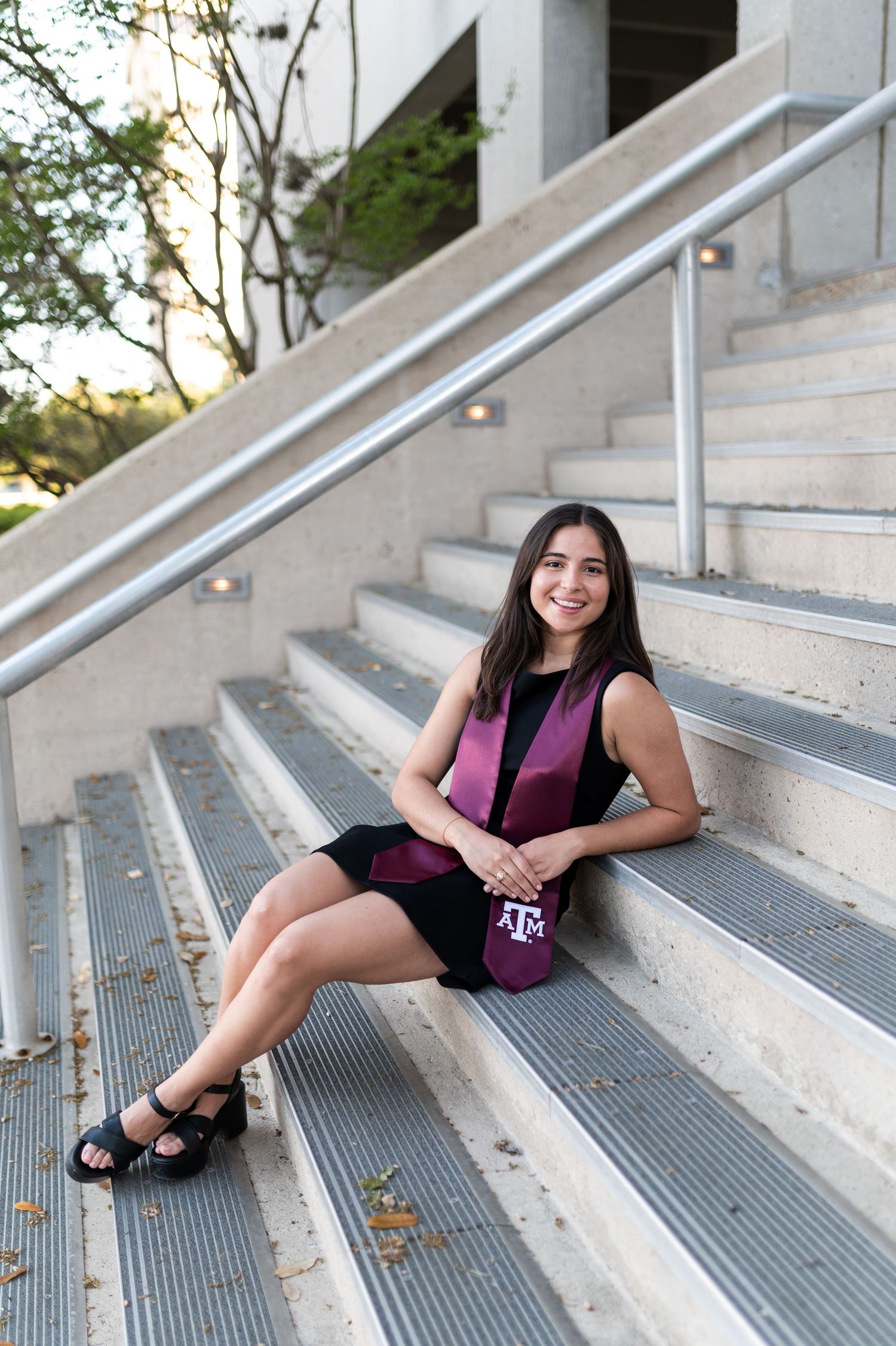 Texas A&M graduate in black dress and maroon stole sits smiling on outdoor metal staircase steps on campus.