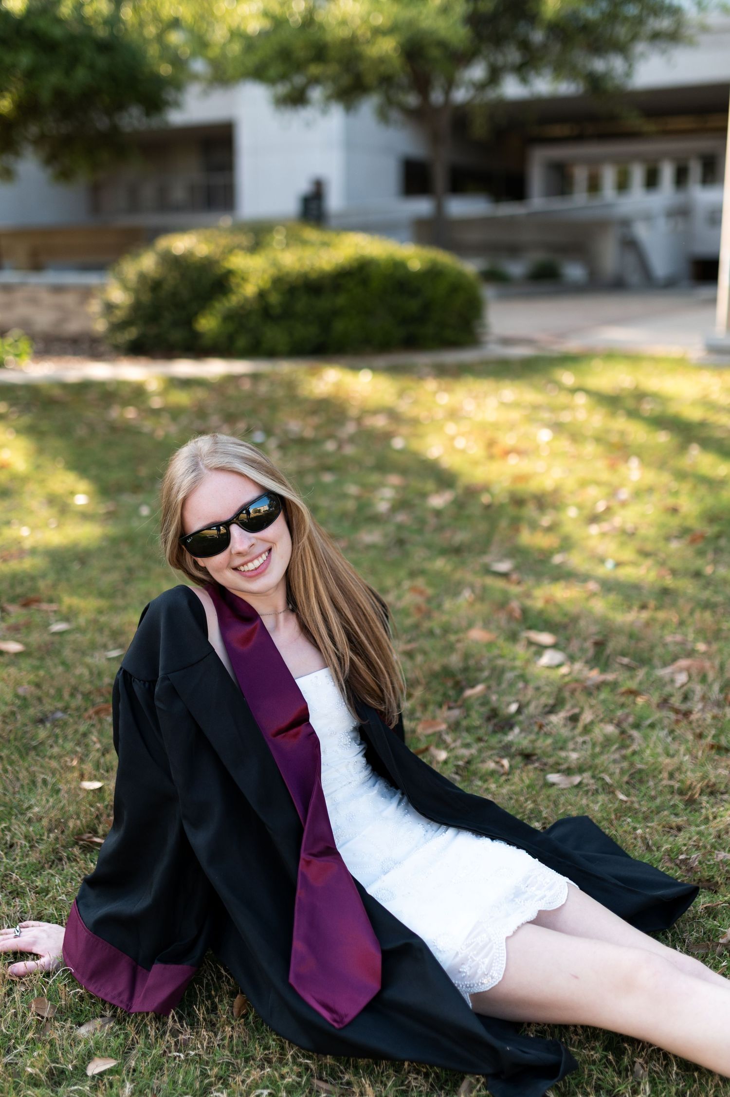 Texas A&M graduate in black gown and maroon stole sits smiling on grass wearing sunglasses outdoors.