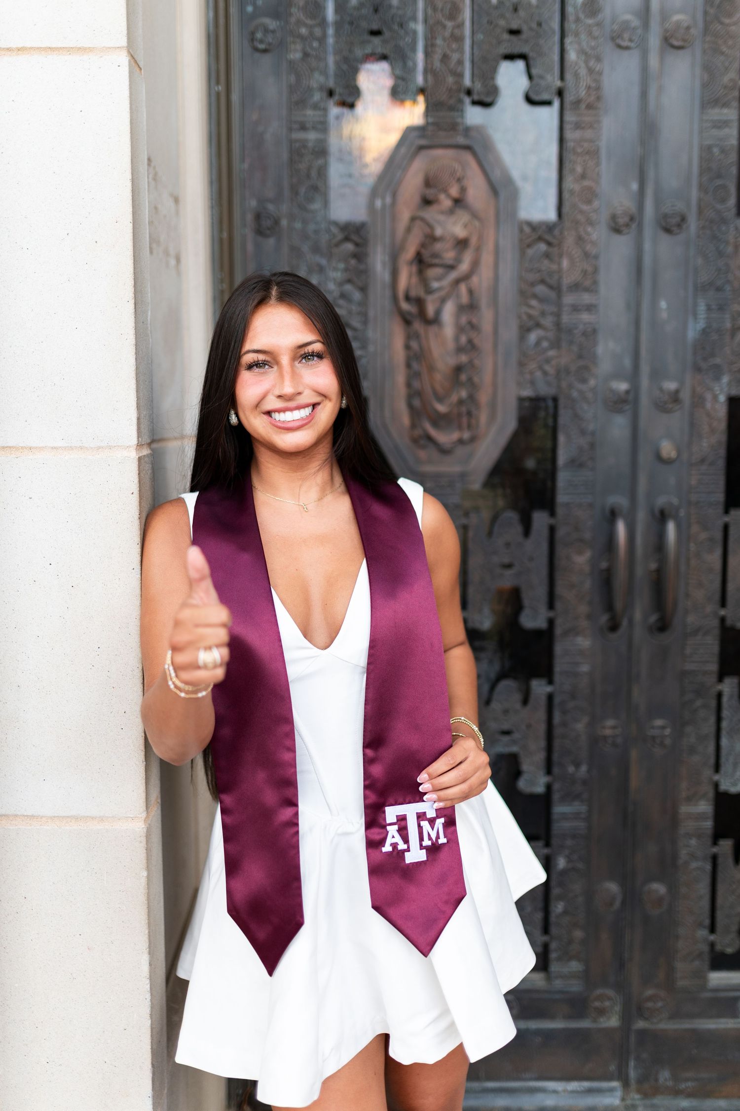 Smiling Texas A&M graduate in white dress and maroon ATM stole gives thumbs up beside ornate bronze campus door. Grad Photos at the Admin