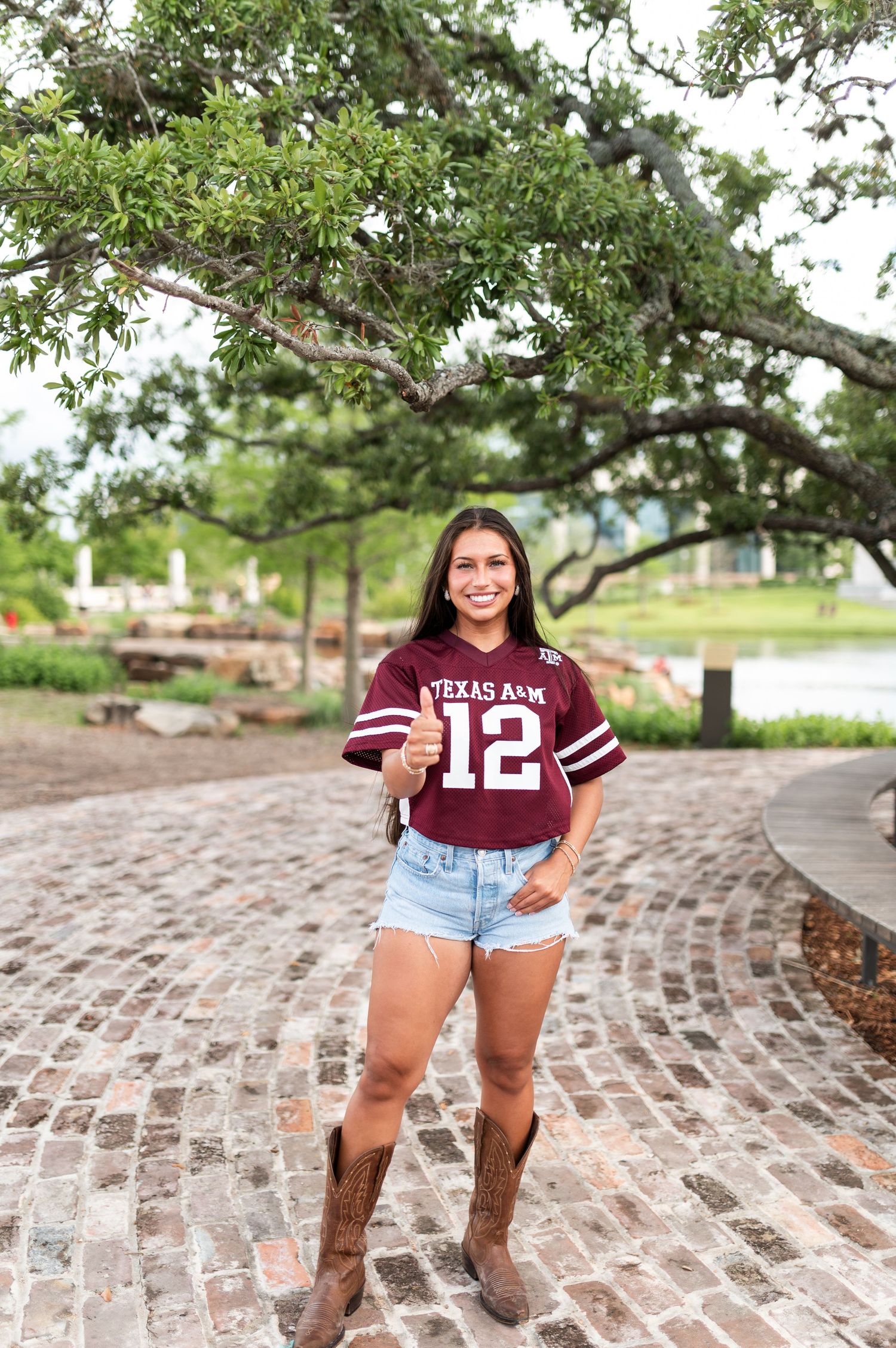 Smiling woman in Texas A&M #12 jersey, denim shorts, and cowboy boots standing on brick path.