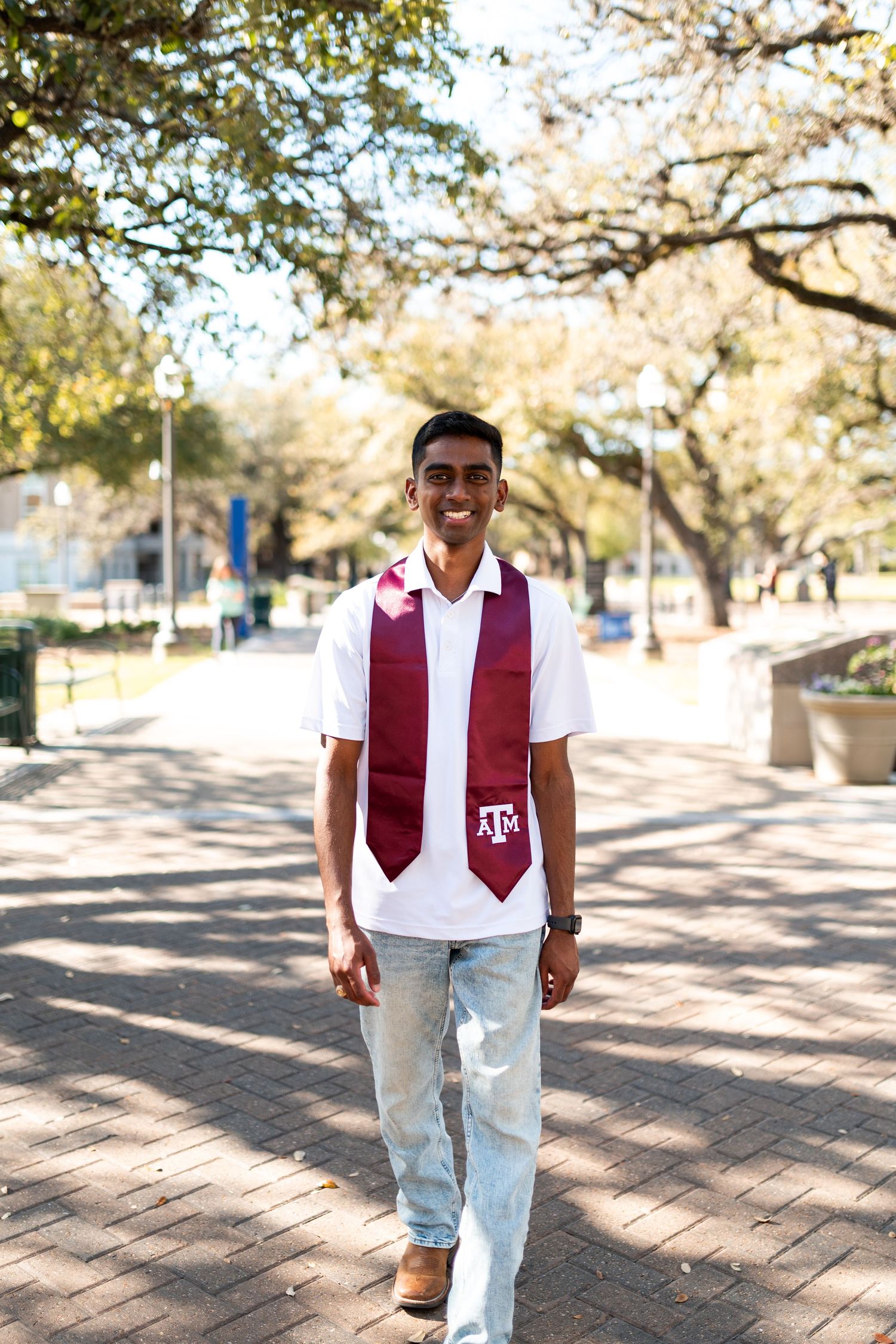 Smiling Texas A&M graduate walks along brick campus path wearing white shirt and maroon graduation stole.