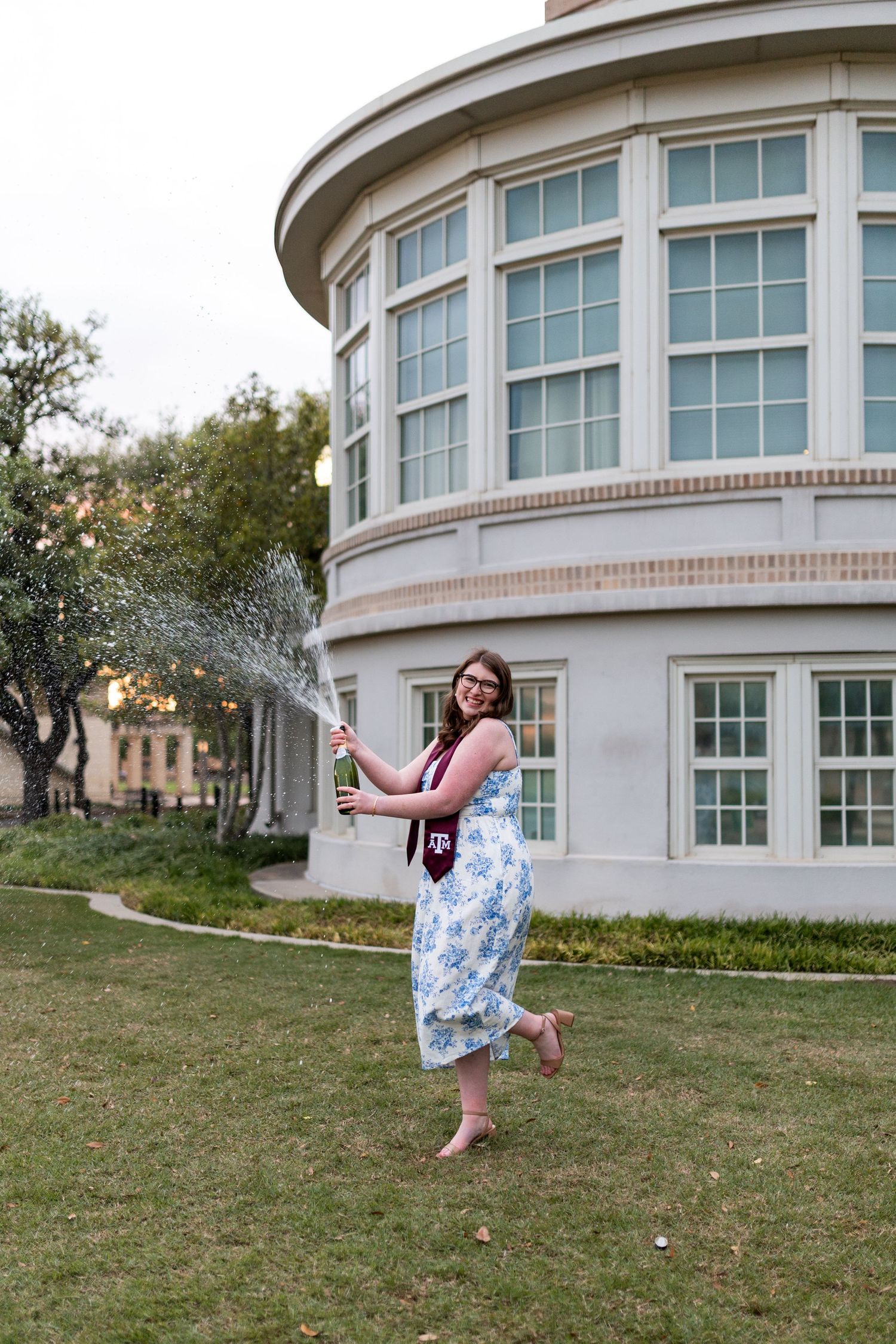 Texas A&M graduate in floral dress joyfully spraying champagne on campus lawn near building.