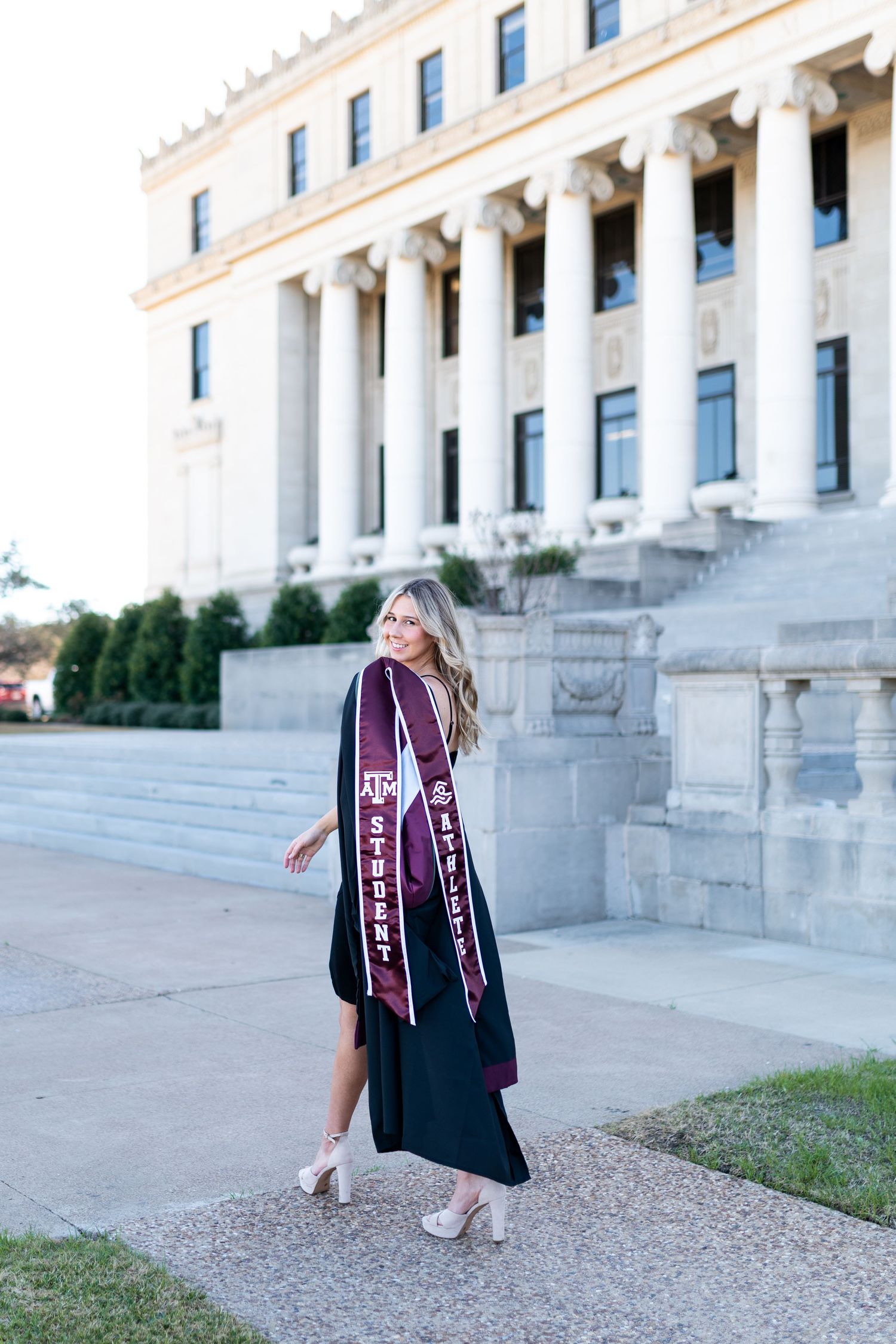 Blonde woman in black dress with Texas A&M graduation stole walking in front of university building. Photos at the Admin building