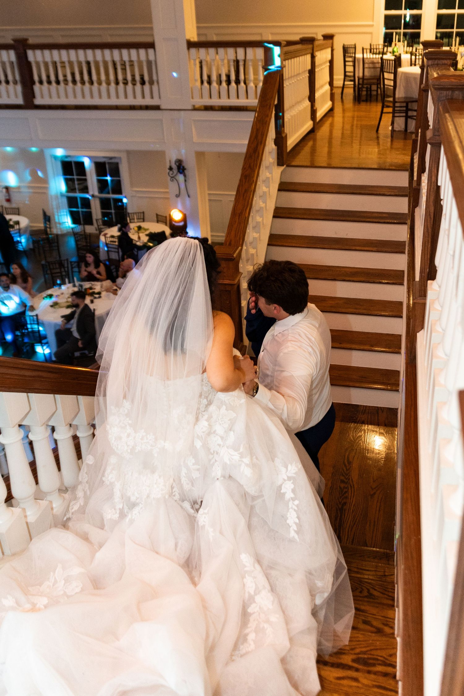 Bride in lace wedding gown and veil ascending grand staircase with helper at elegant wedding reception venue.