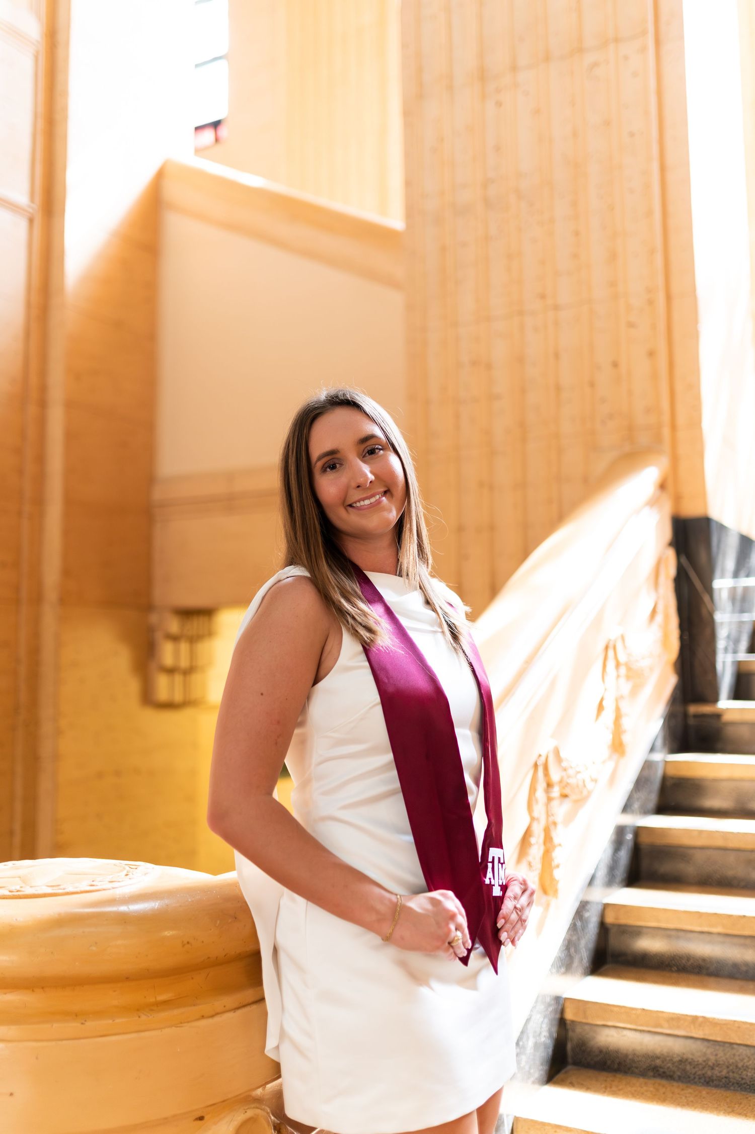 Smiling female graduate in white dress and maroon stole poses on ornate staircase. Grad Photos at the Texas A&M Admin building