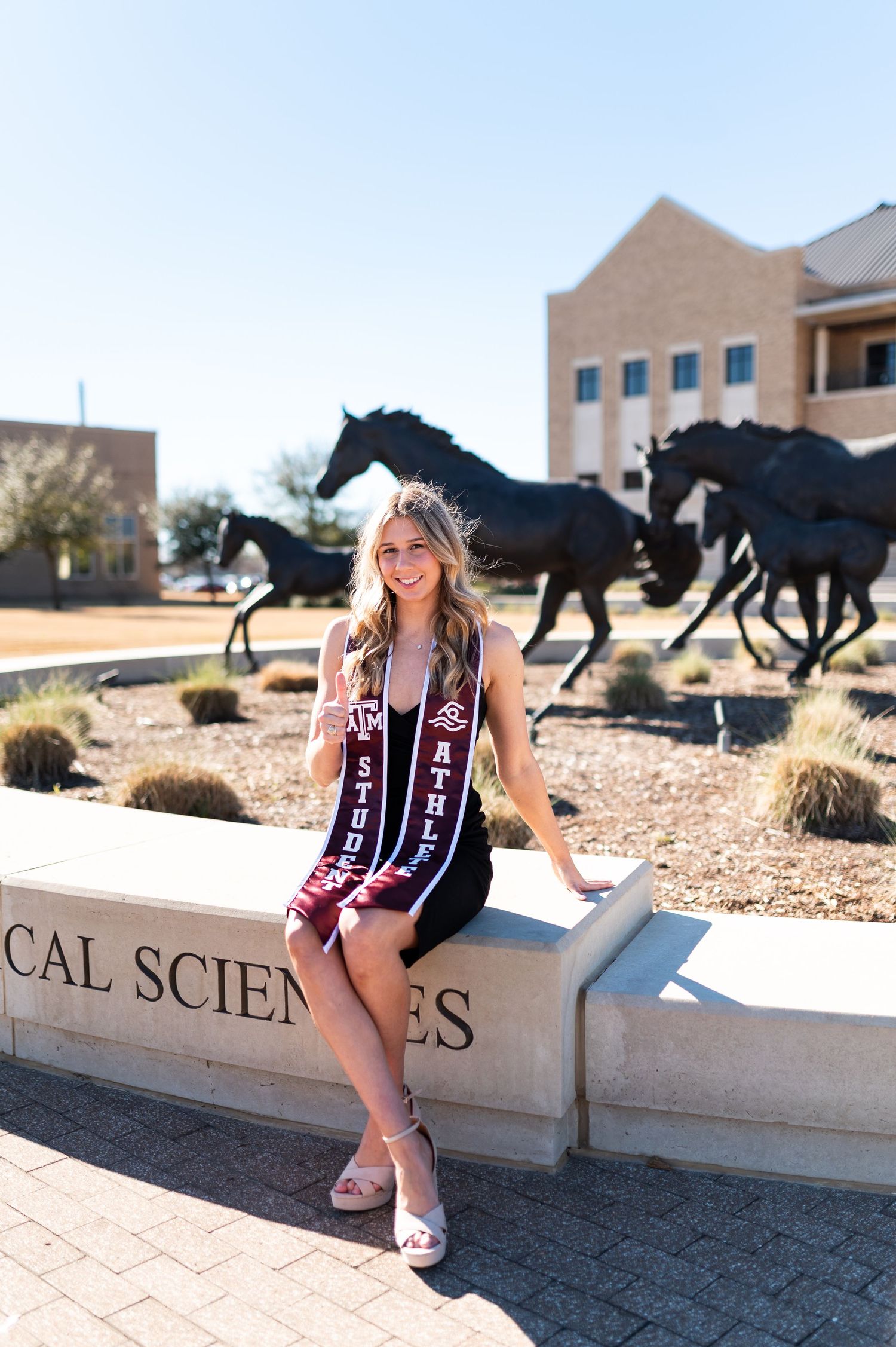 Texas A&M student athlete poses by horse sculptures wearing maroon Student Athlete sash outdoors.